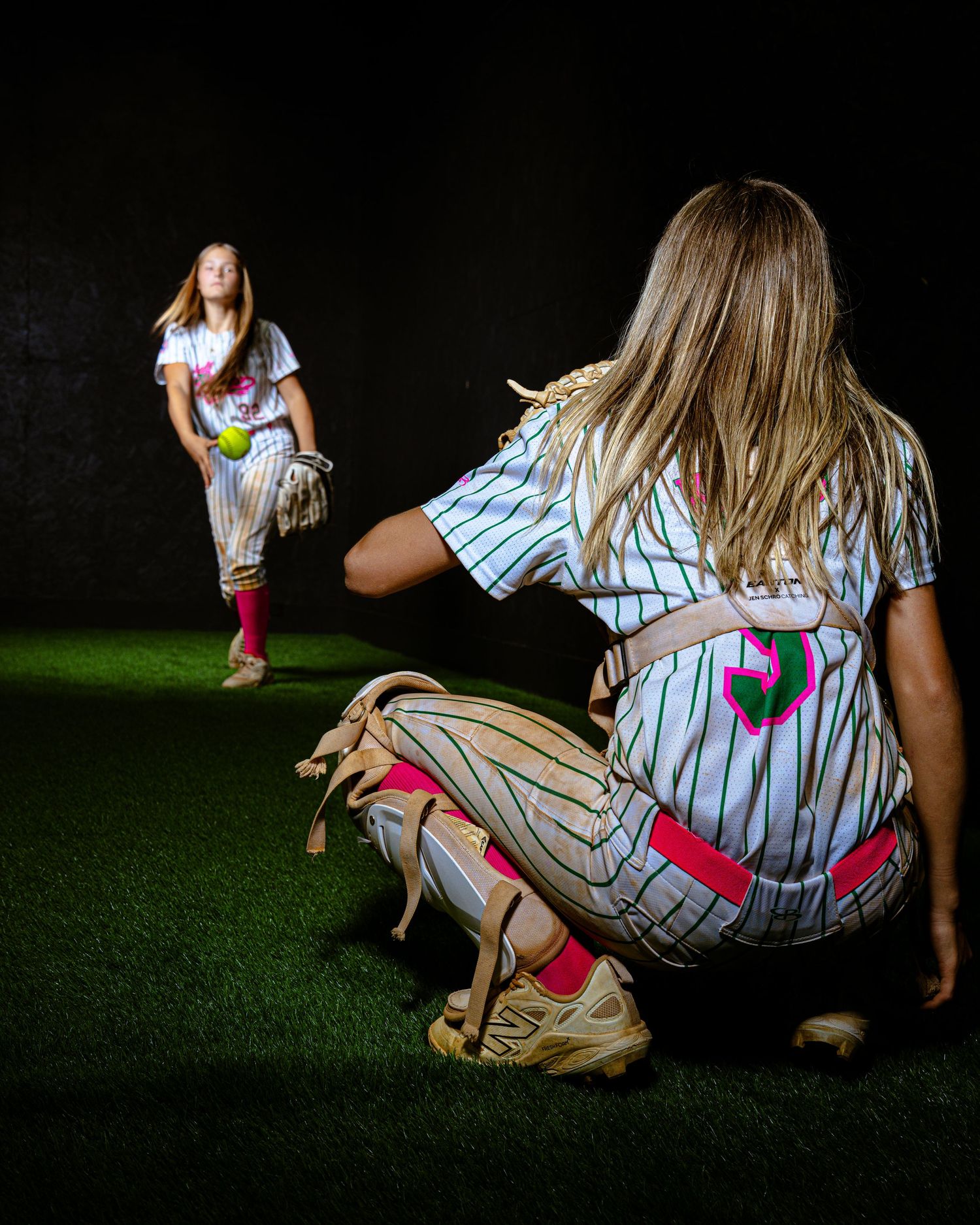 Two softball players in pinstripe uniforms practice pitching and catching on green turf against a dark background.