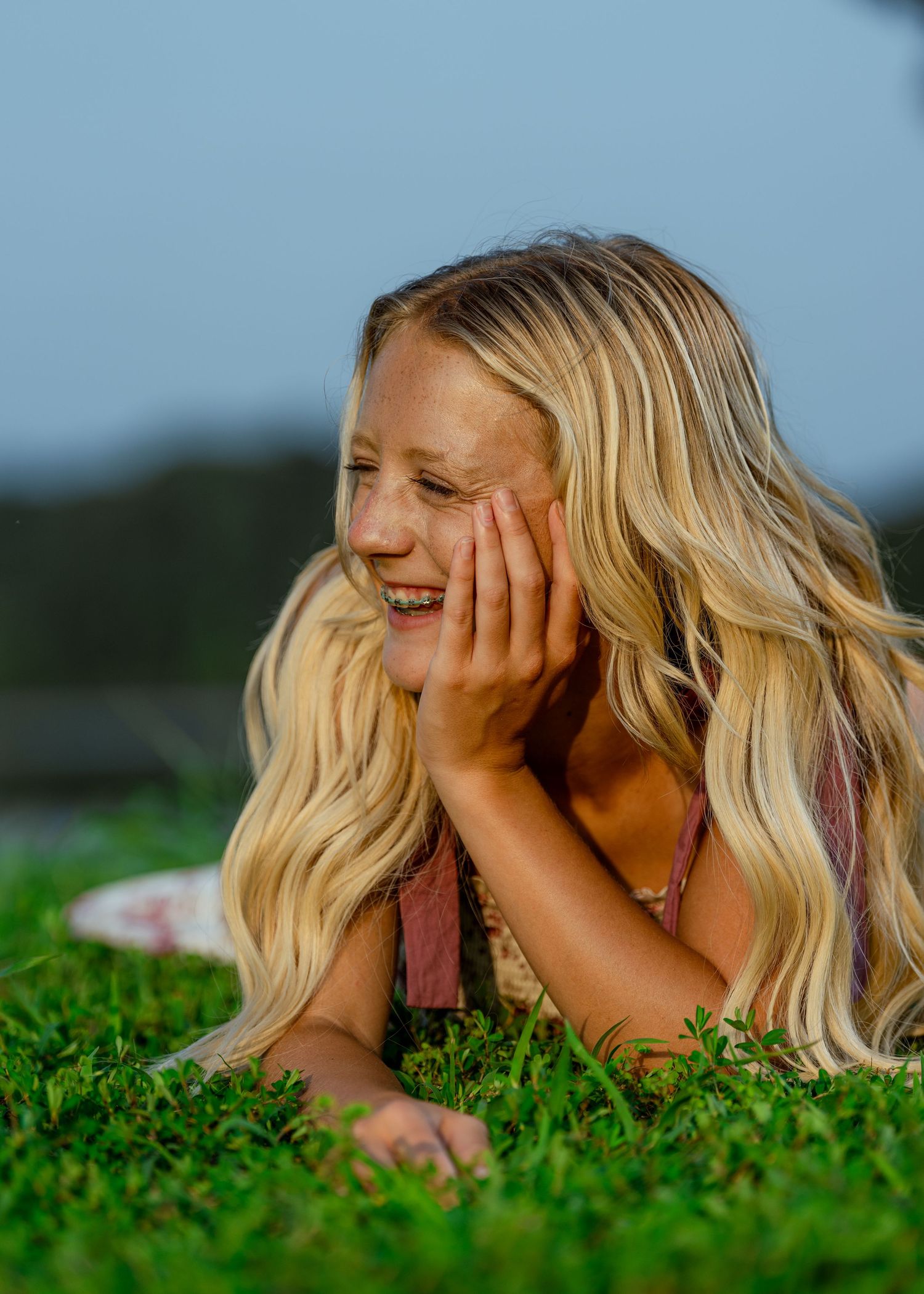 A smiling blonde girl with braces lies on green grass, resting her chin on her hand on a sunny day.