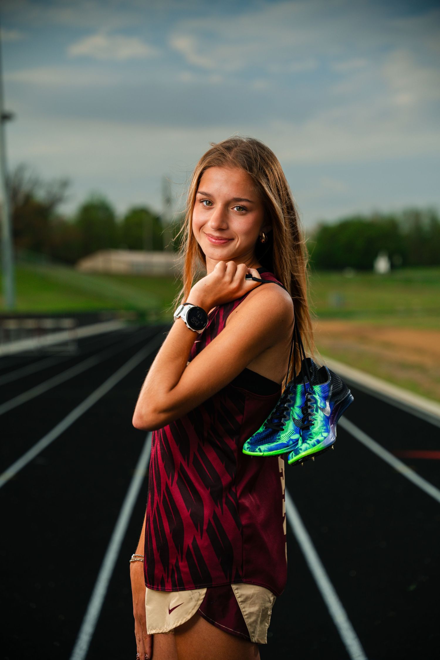 Young female track athlete holding colorful running spikes on a black running track, wearing a red tie-dye tank top.