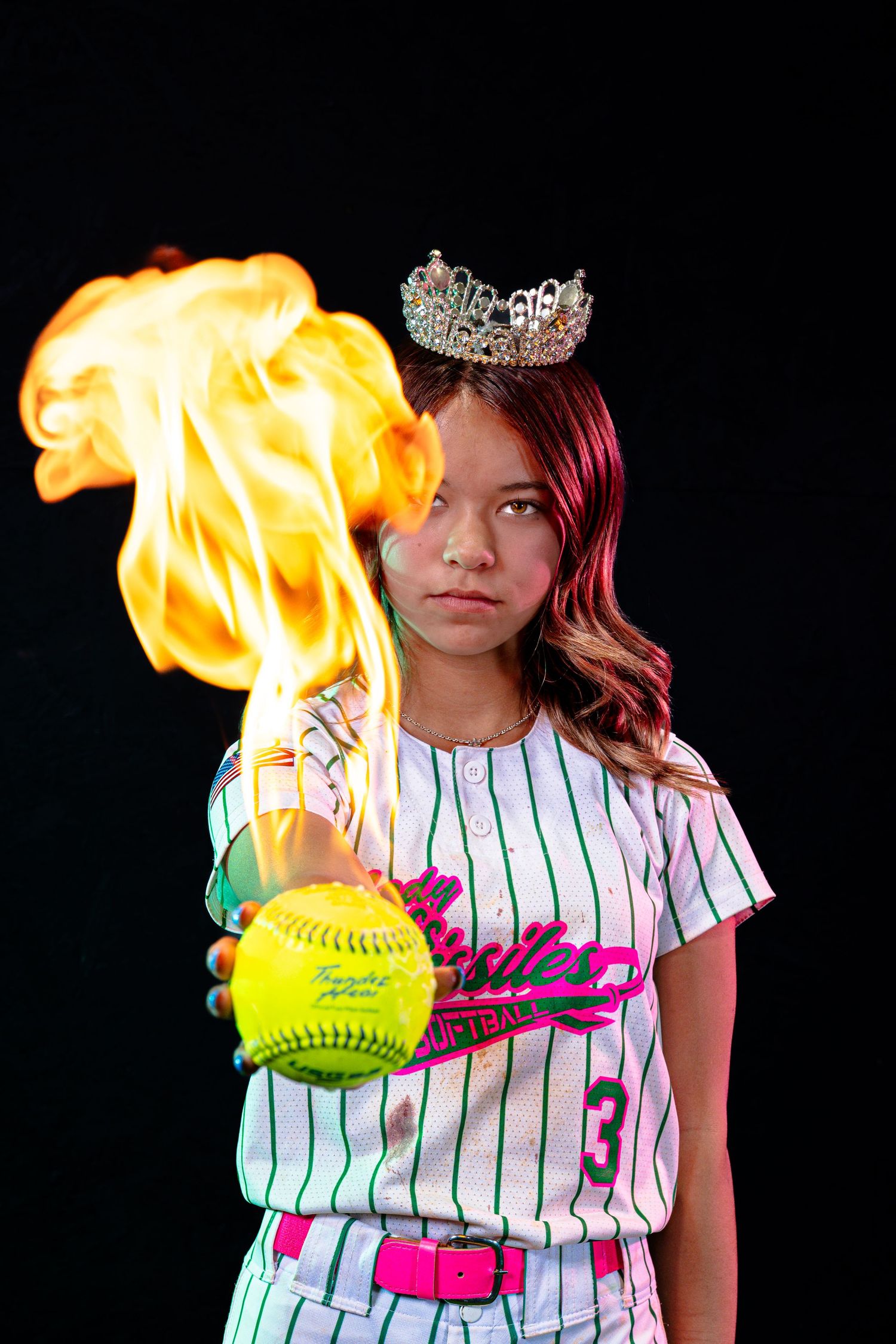 Young girl in softball uniform and tiara holds a flaming yellow softball against a black background.
