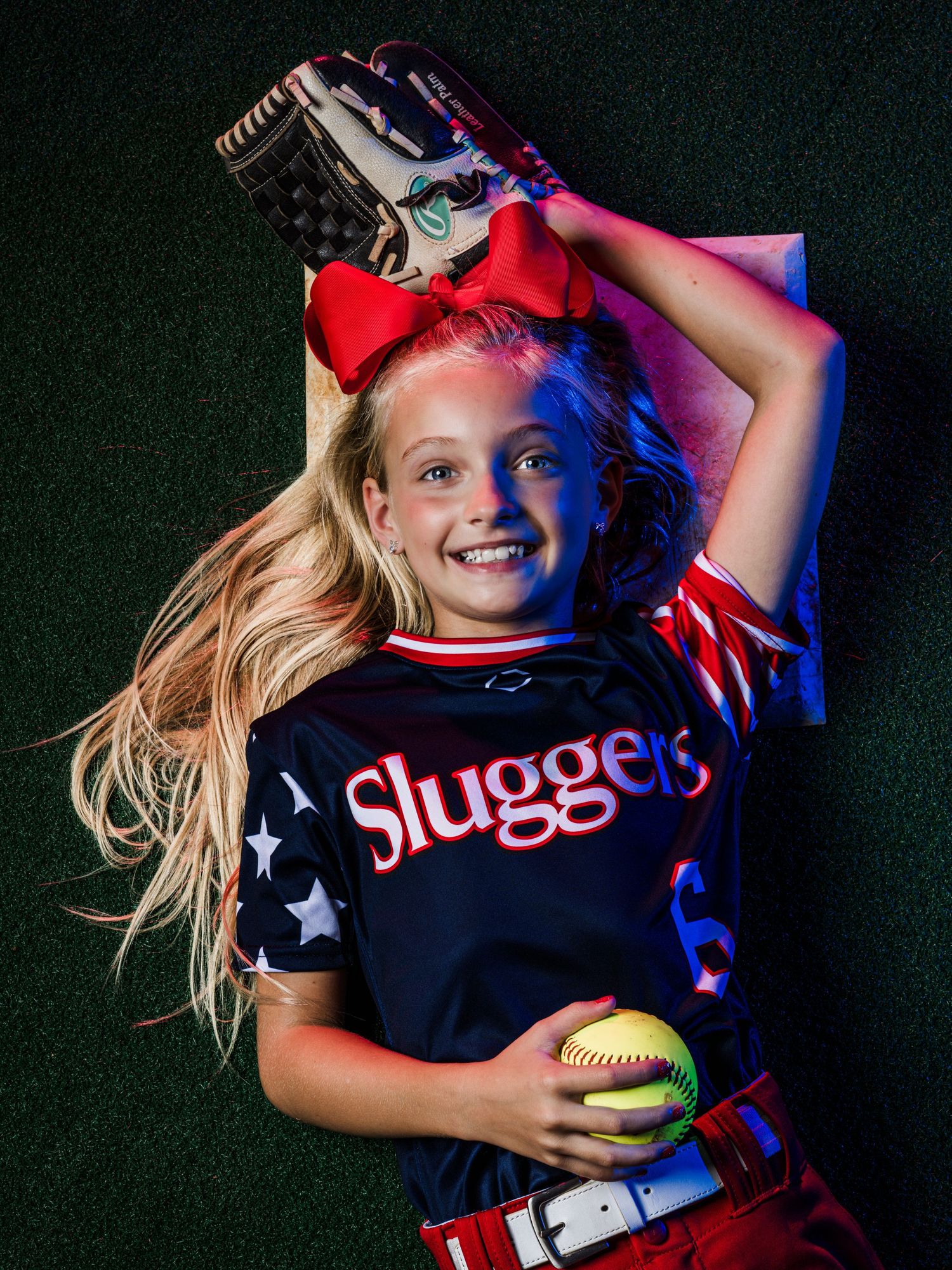 Young softball player in Sluggers jersey lying on field, holding ball and glove, wearing red bow in hair.