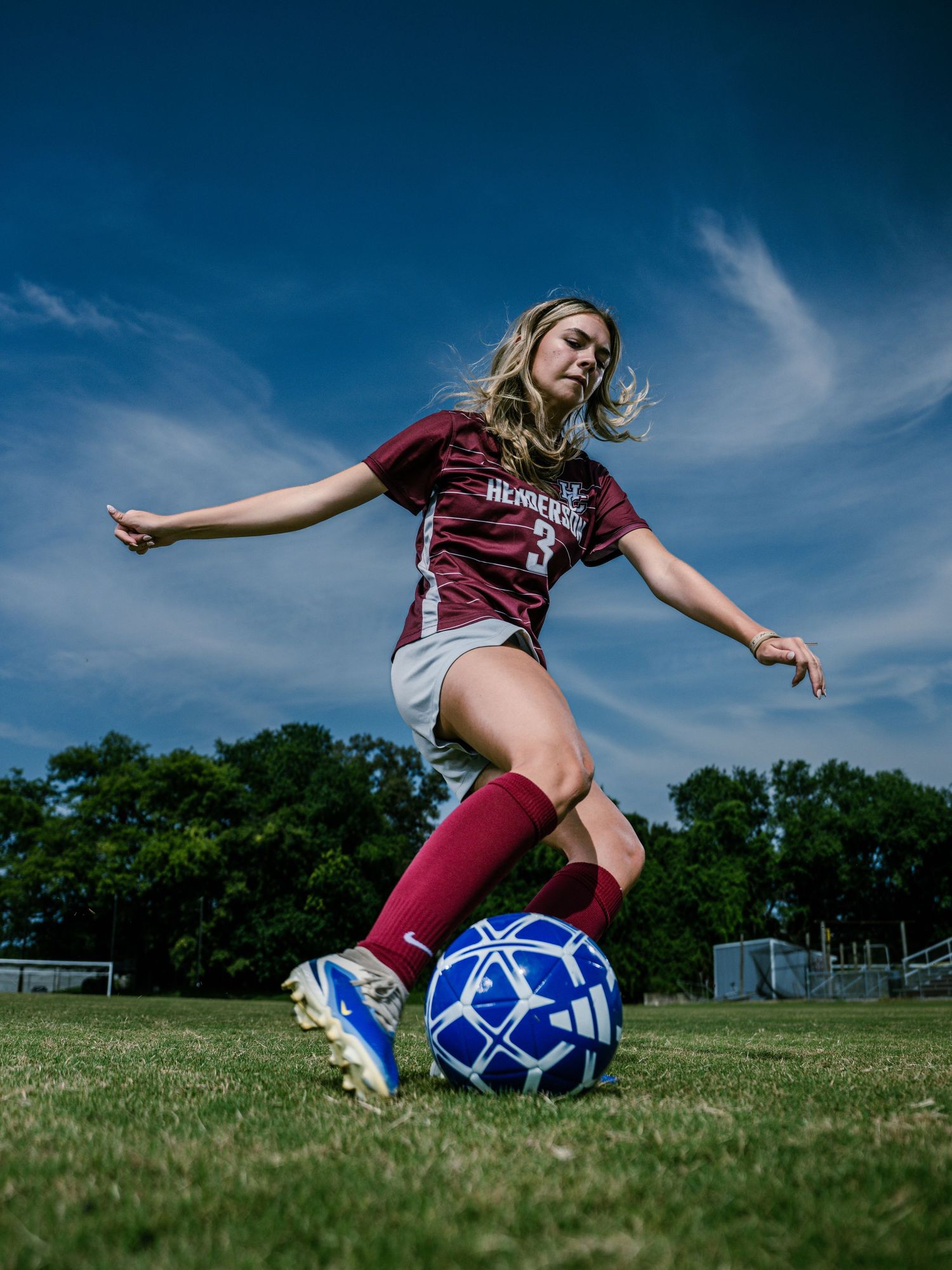 Female soccer player in maroon jersey kicking a blue ball on grass field under blue sky.