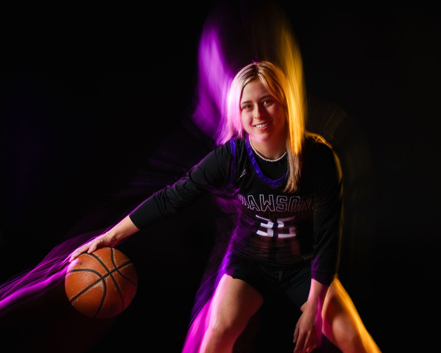 Female basketball player in dark jersey poses with ball under colorful neon lights against black background.