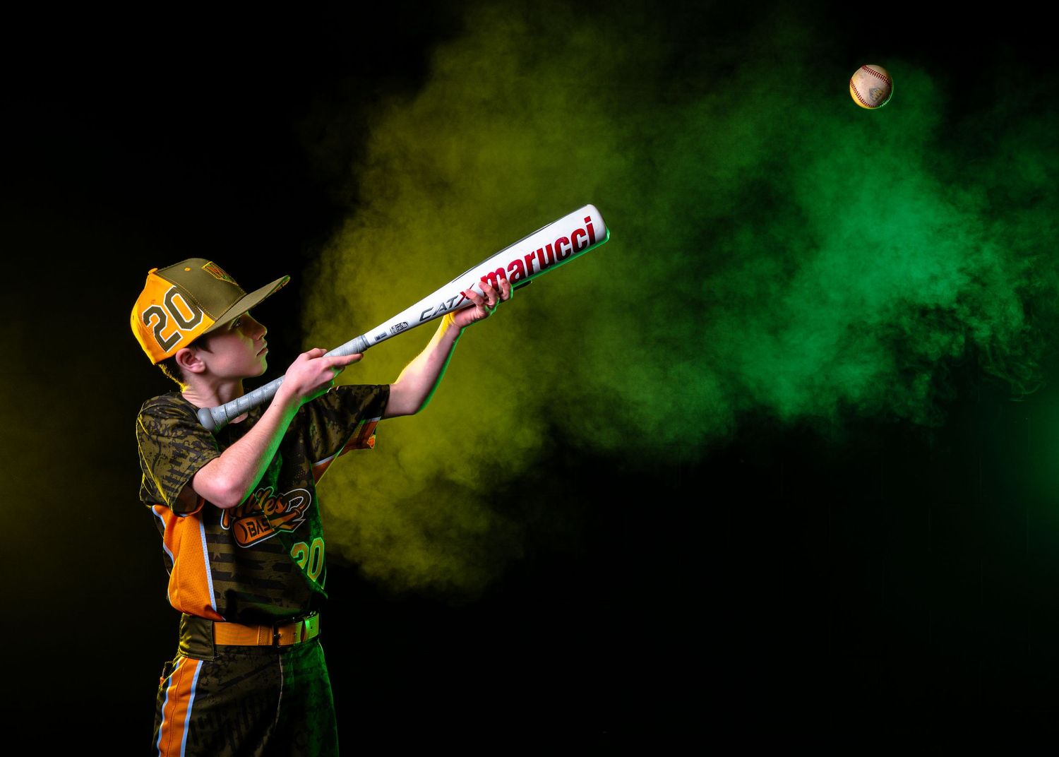 Youth baseball player swinging a Marucci bat with colorful green and yellow powder explosion on dark background.