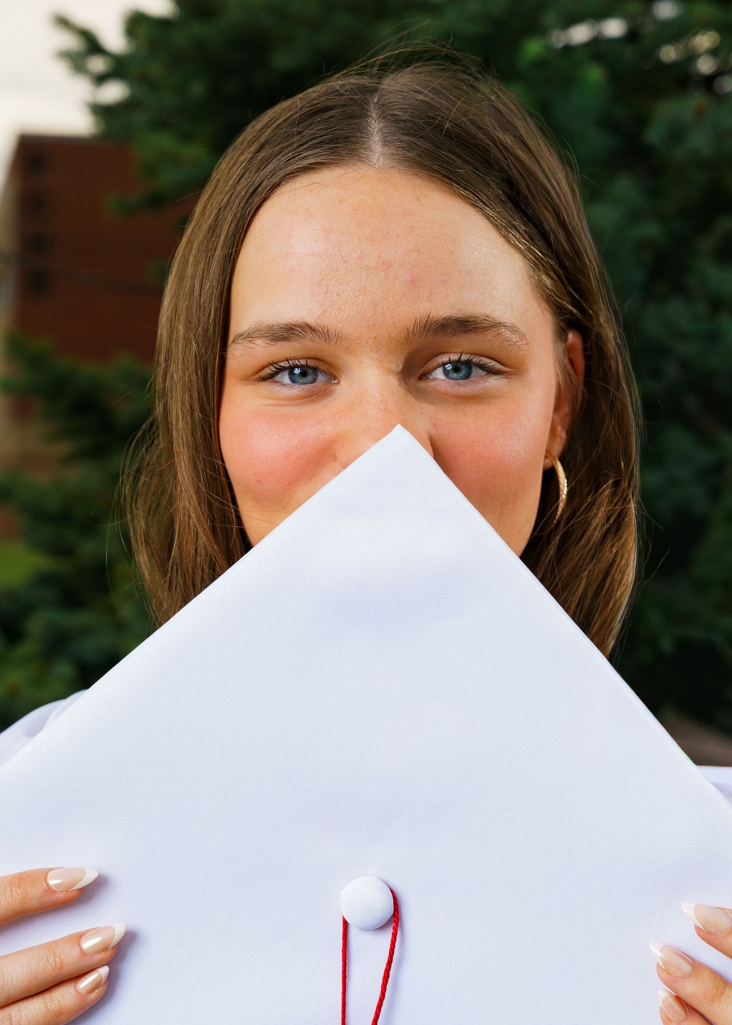 Young woman holding white graduation cap in front of her face, showing blue eyes and brown hair outdoors.