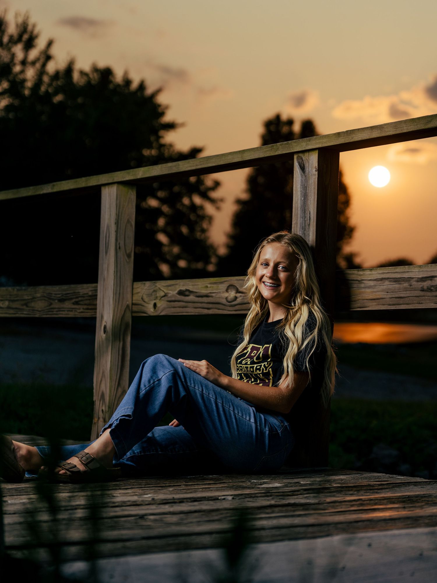 Young blonde woman smiling on wooden dock at golden sunset, wearing graphic tee and jeans.