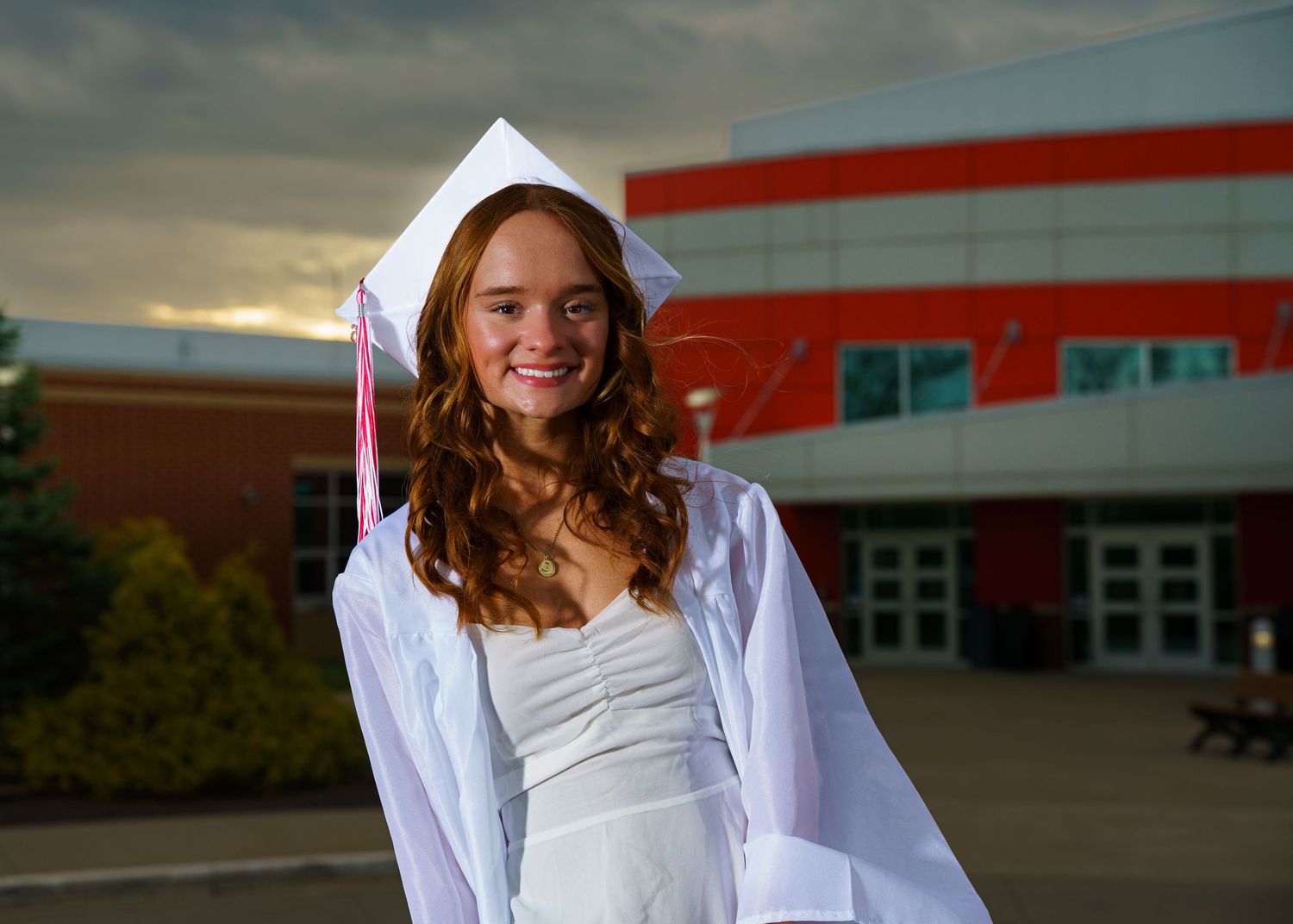 Smiling teenage girl in white graduation cap and gown posing outdoors in front of a brick school building.