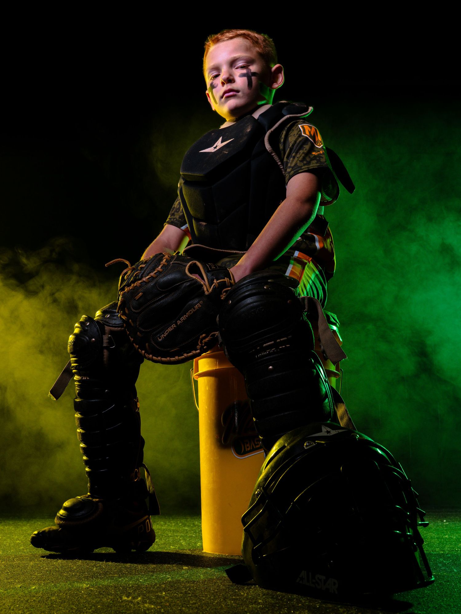 Young baseball catcher in full gear poses dramatically with green smoke and dark background.