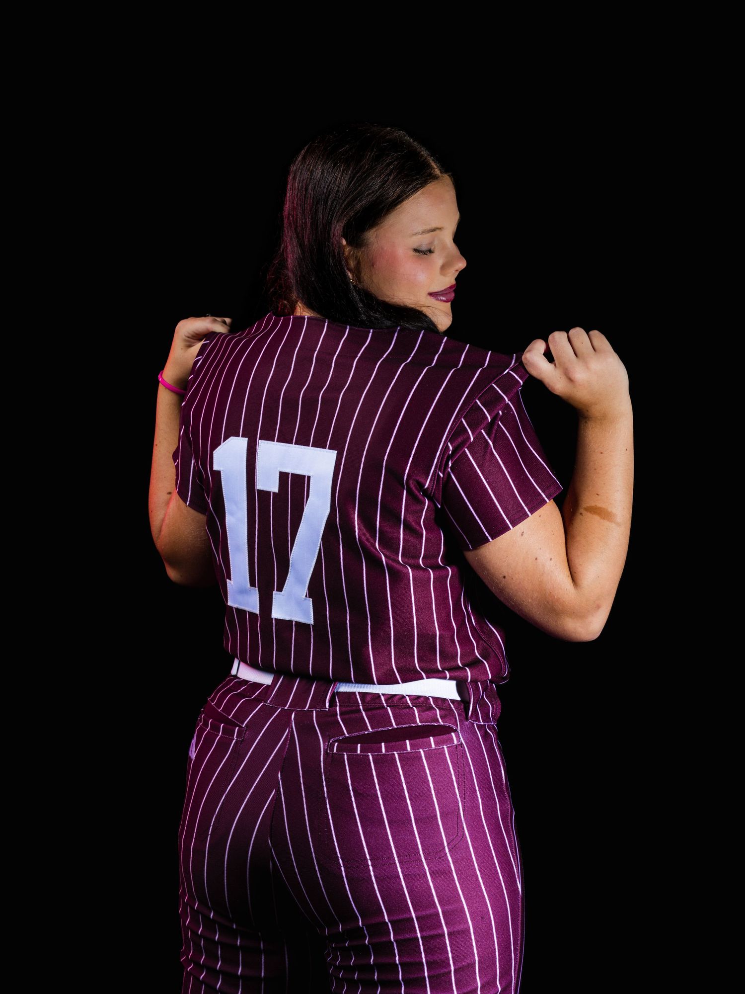 Woman wearing maroon pinstripe softball uniform number 17, posed with back to camera against black background.