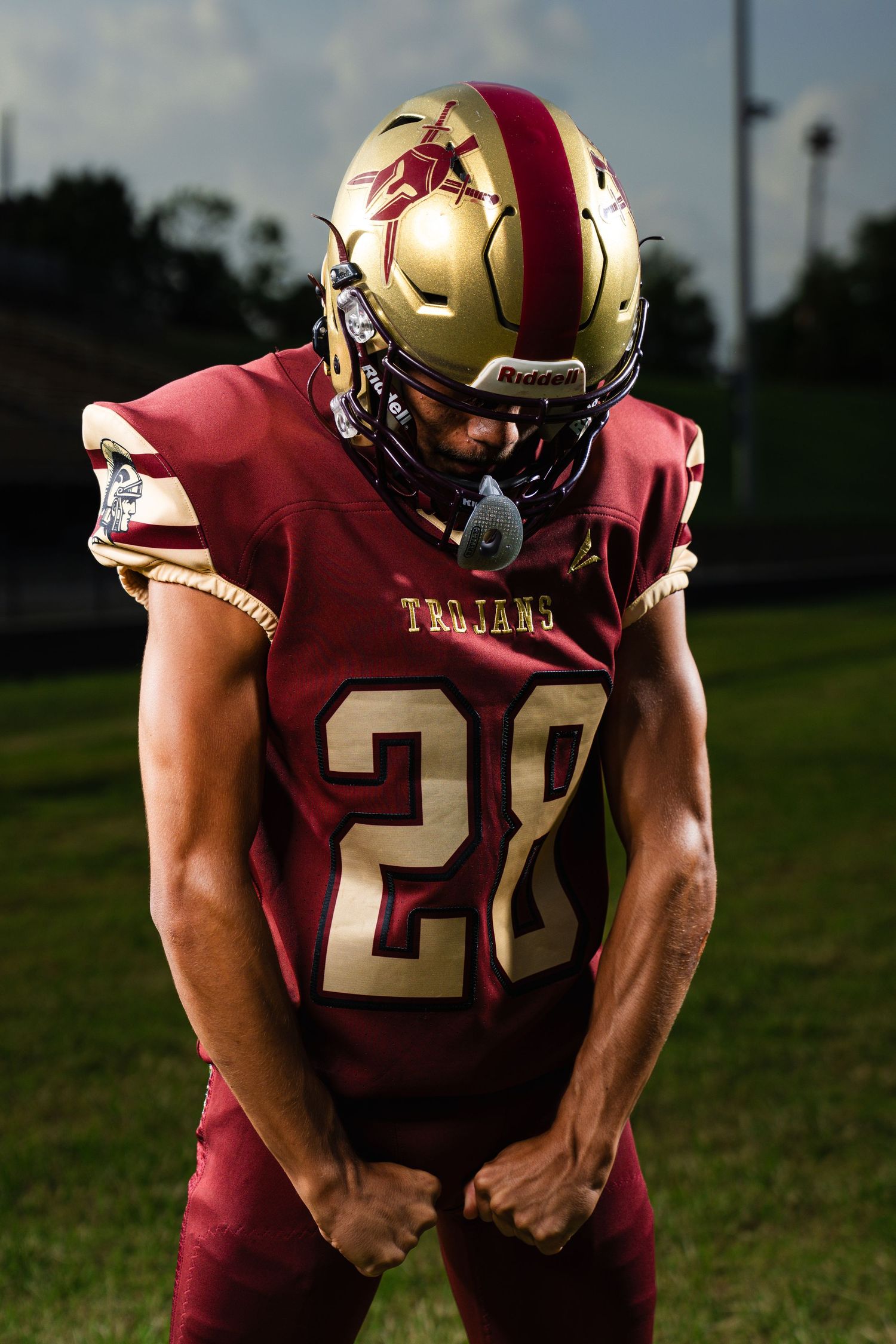 Football player wearing Trojans #28 maroon and gold uniform with Riddell helmet stands on field in dramatic pose.