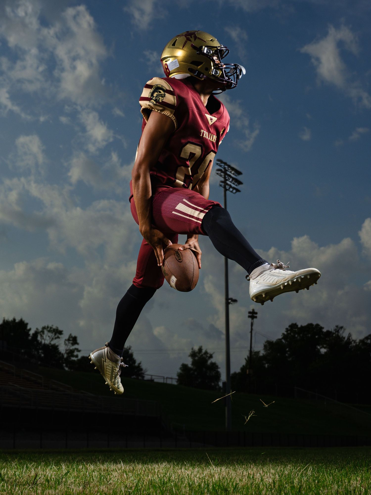 Football player in maroon and gold uniform leaping mid-air on field, holding ball, wearing gold helmet under blue sky.