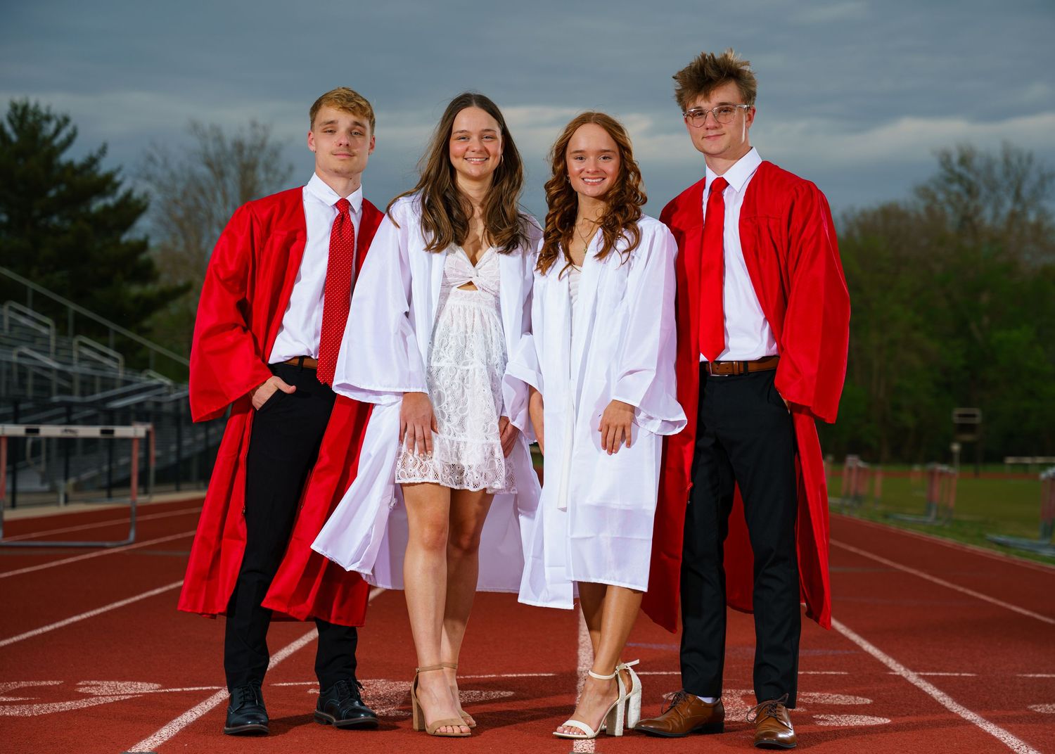 Four high school graduates in red and white caps and gowns posing on a track field at graduation.