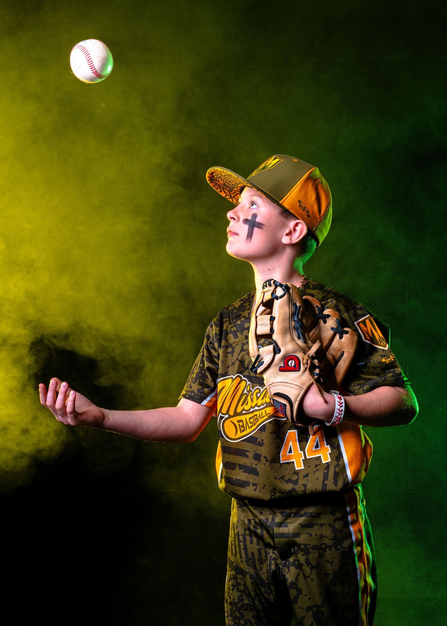 Young baseball player in camo uniform #44 tossing a ball in dramatic green and yellow smoke.