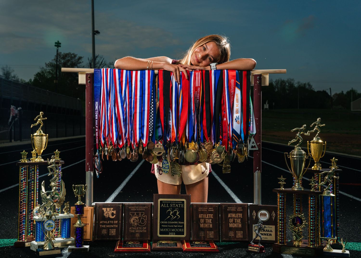 Young female athlete posing proudly with numerous medals, ribbons, and trophies displayed on a rack at a track field.