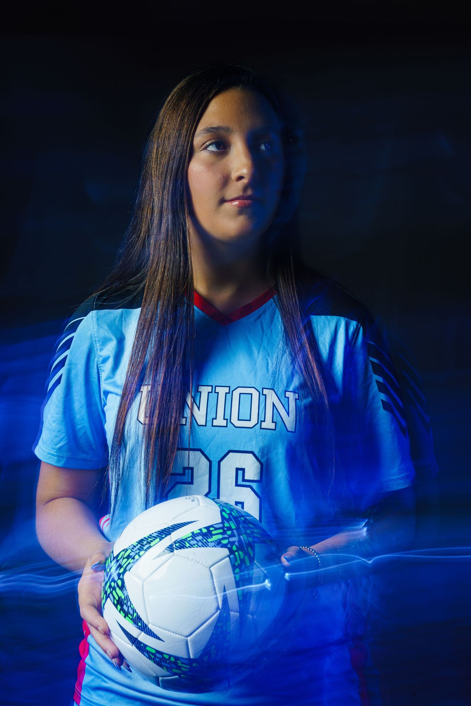 Young female soccer player in Union #26 blue jersey holds a soccer ball, posed against a dark background with blue light effects.