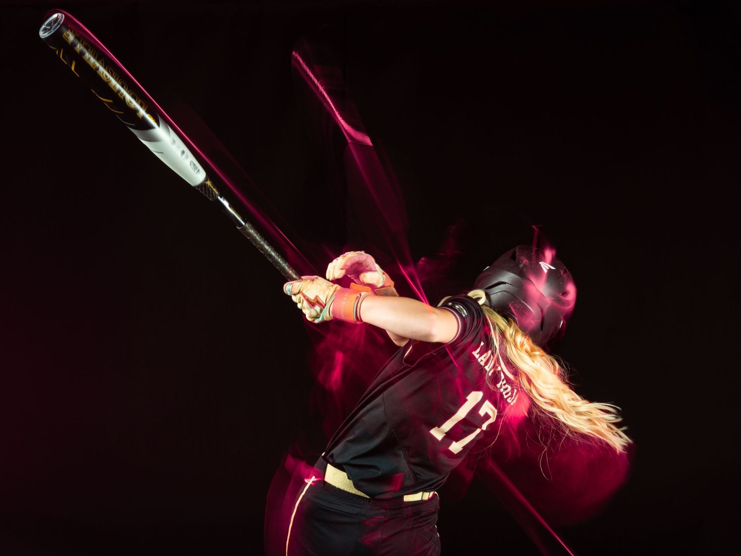 Female softball player in maroon jersey number 11 swings bat with dynamic motion blur effect on dark background.