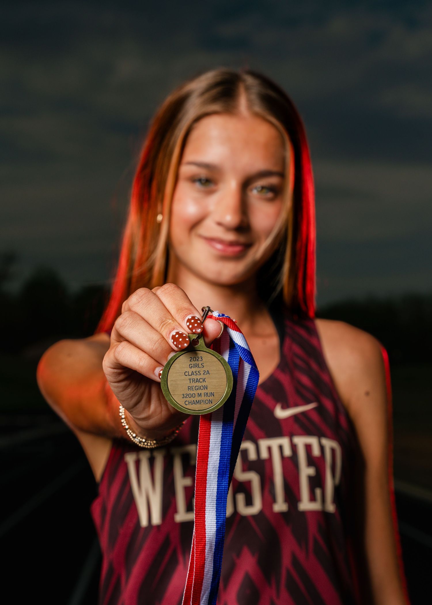 Young female athlete in Webster jersey proudly holds a gold medal toward camera on a track field.