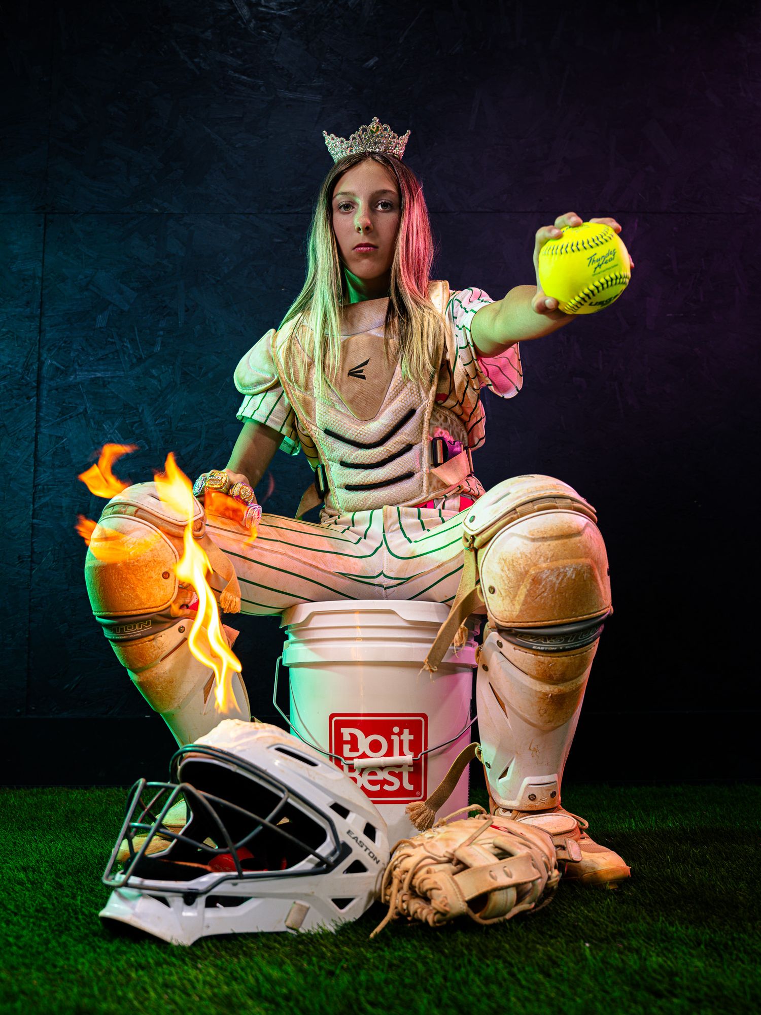 Young softball catcher in gear and crown holds ball, sits on bucket with flaming glove, helmet and mitt on turf.