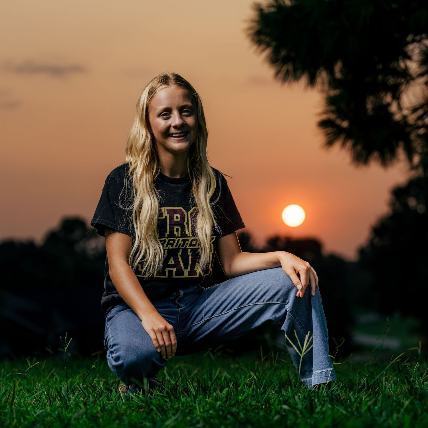 Young blonde woman smiling in black graphic tee and jeans, sitting on grass during a golden sunset outdoors.