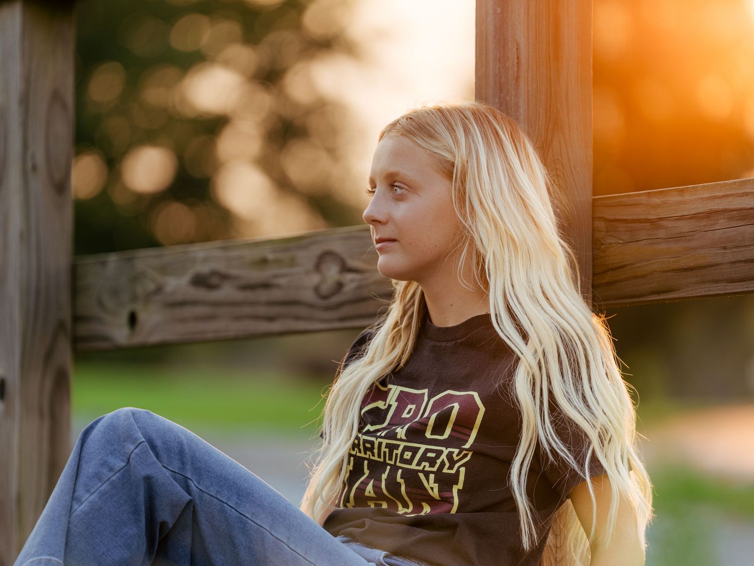 Young blonde woman in brown graphic tee and jeans relaxing outdoors in warm golden sunset light.