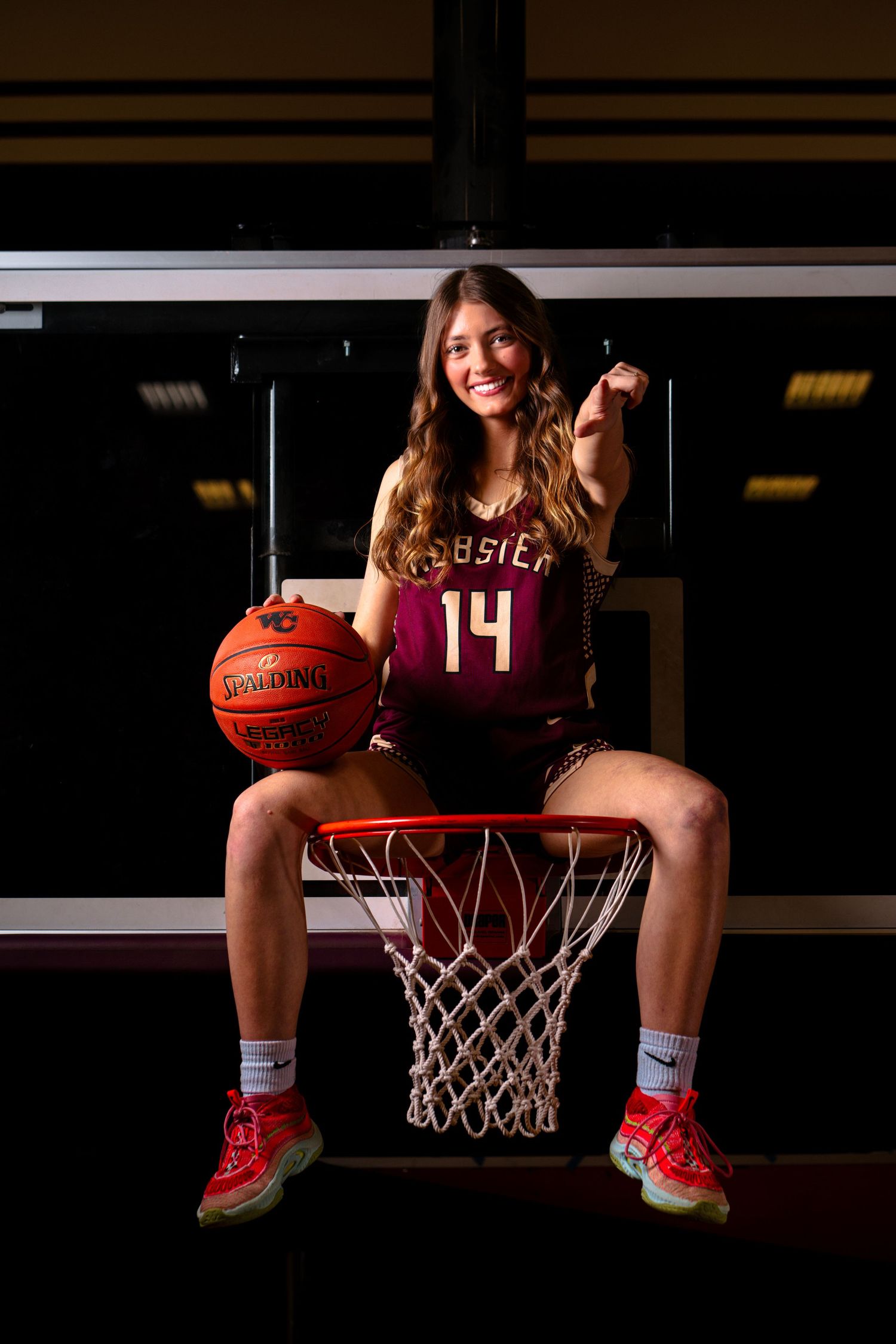 Female basketball player #14 sits smiling on a hoop rim holding a Spalding ball in a maroon jersey.