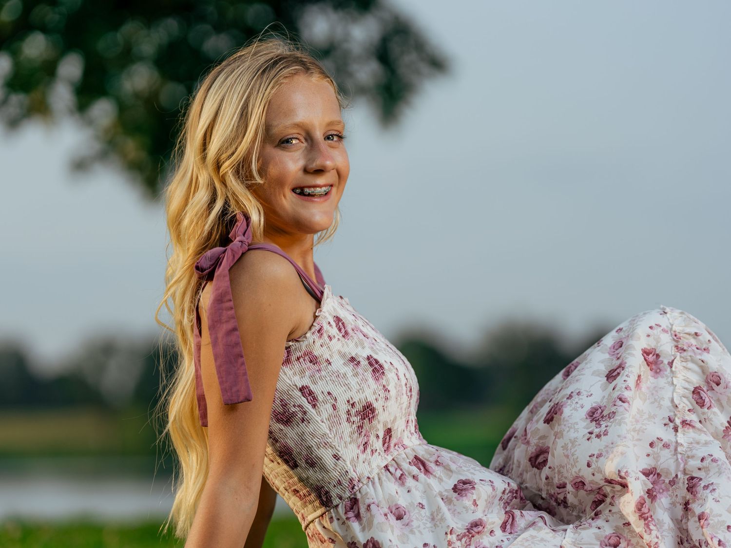 Young blonde woman in floral pink dress smiling outdoors near a lake on a cloudy day.