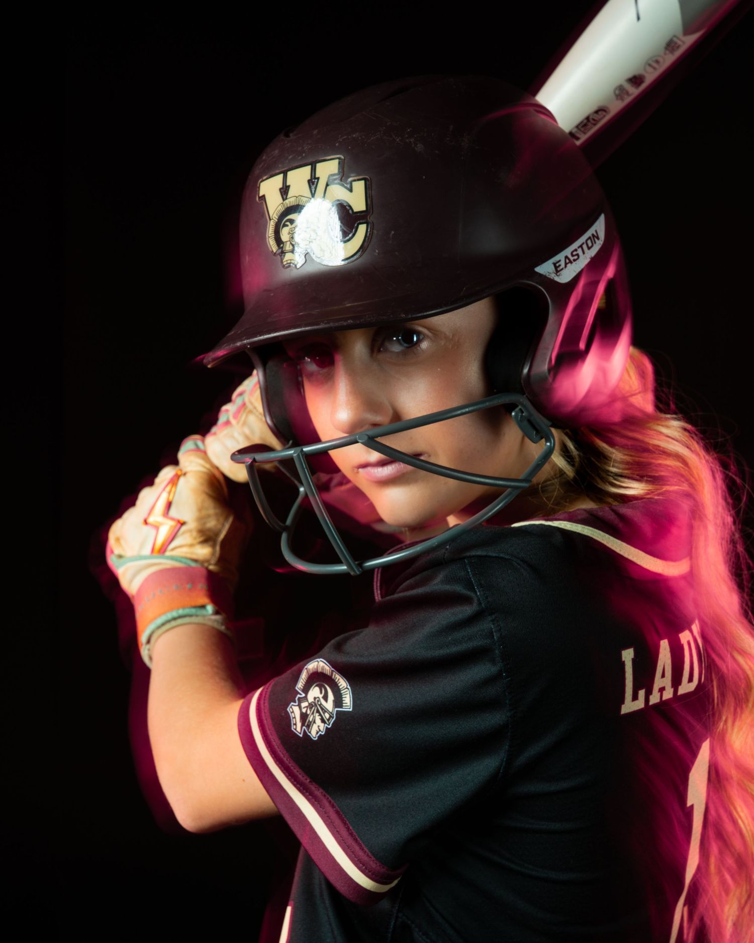 Female softball player in maroon uniform and helmet holds bat ready to swing in dramatic studio portrait.