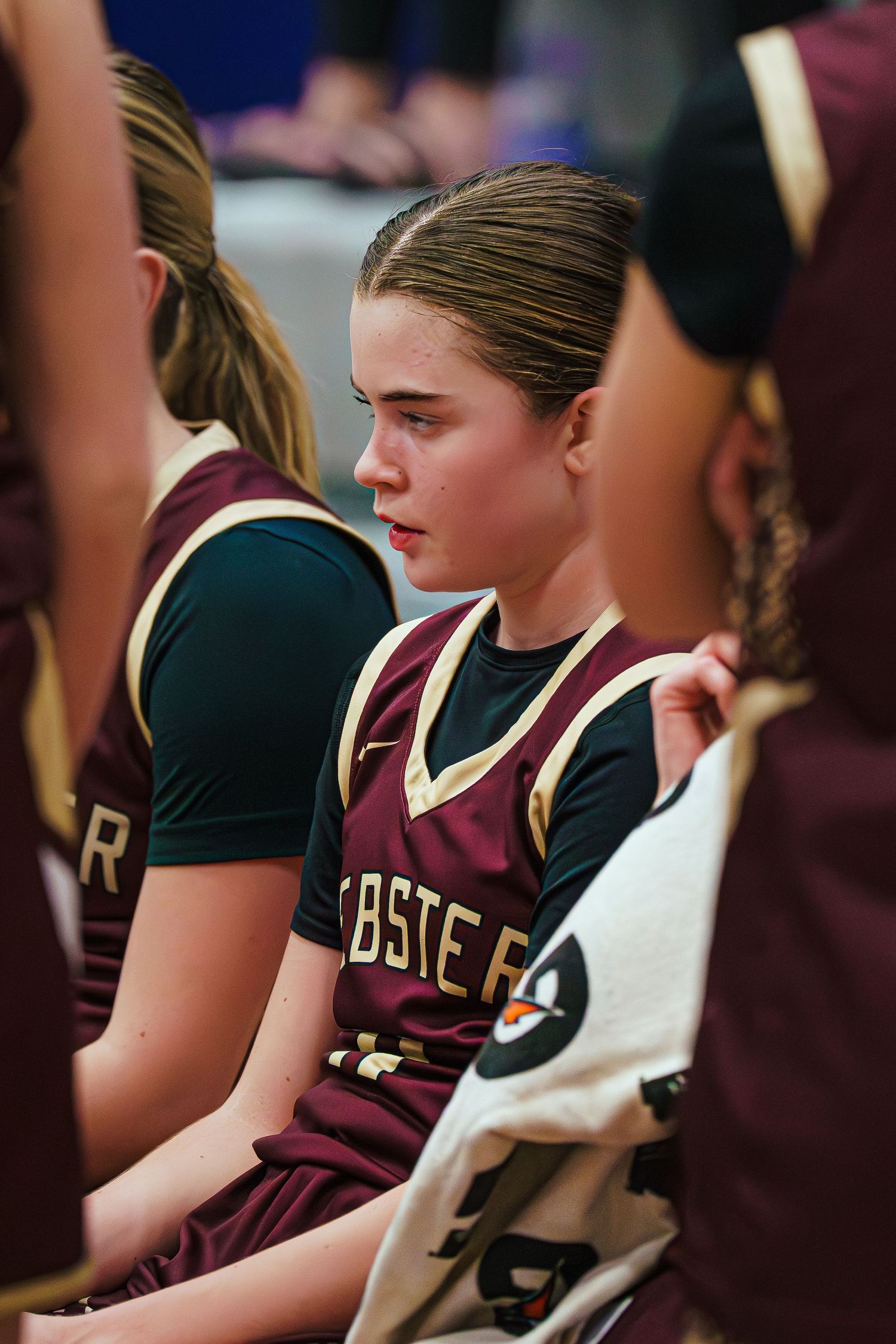 Young female basketball player in maroon jersey sits focused on the bench during a game timeout.