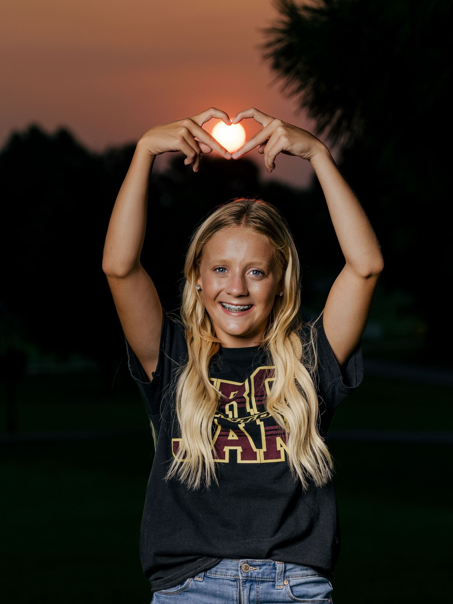 Smiling blonde teen girl makes heart shape with hands above head at sunset wearing a black graphic tee.