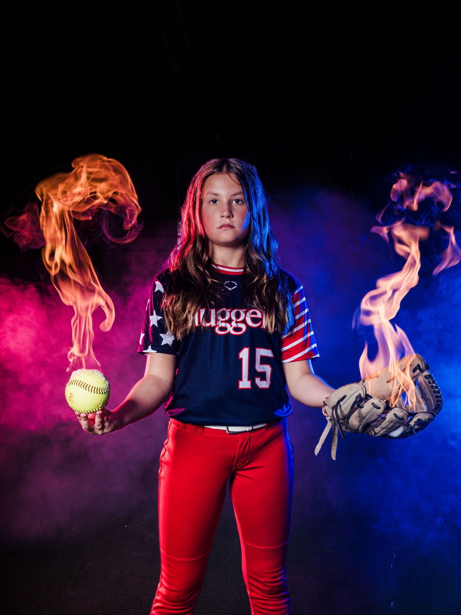 Young softball player #15 in Nuggets jersey holds flaming ball and glove with dramatic blue and red smoke effects.