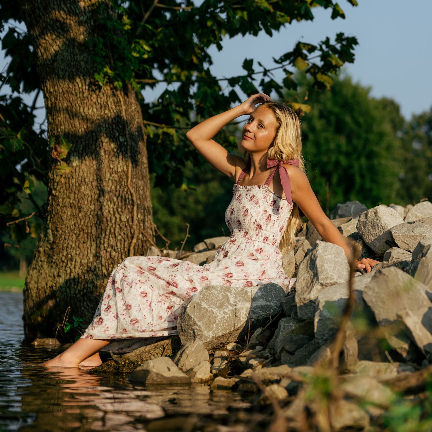 Blonde woman in floral sundress sitting on rocks by water near a large tree in golden sunlight.