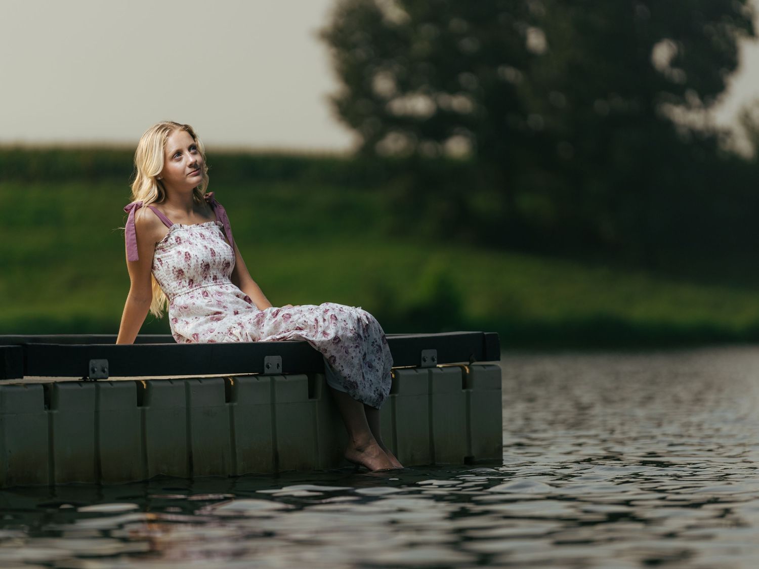 Blonde woman in floral dress sitting on edge of a boat on calm water with green trees in background.