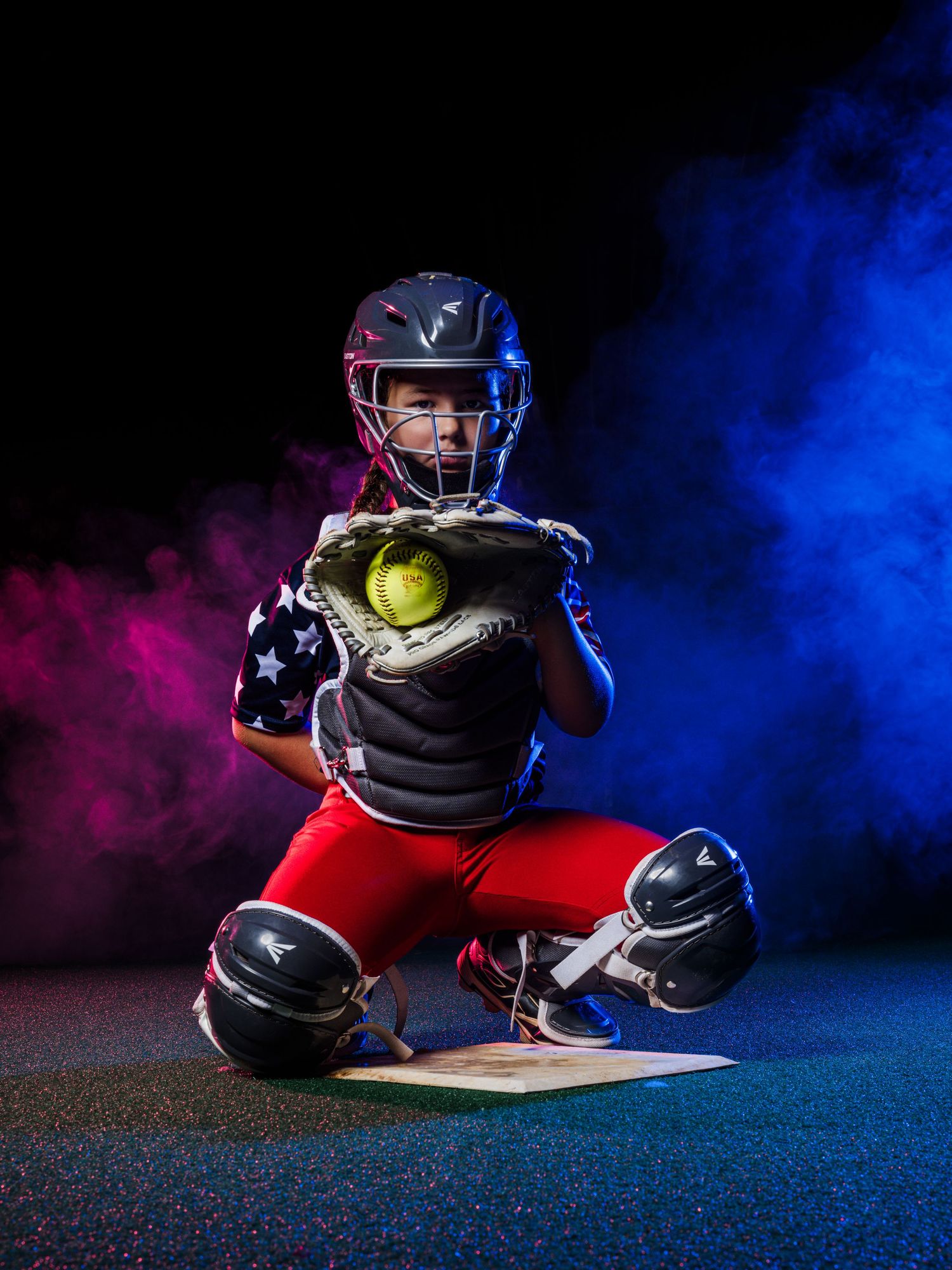 Young softball catcher in full gear crouching in position, wearing helmet and chest protector, catching a yellow ball.