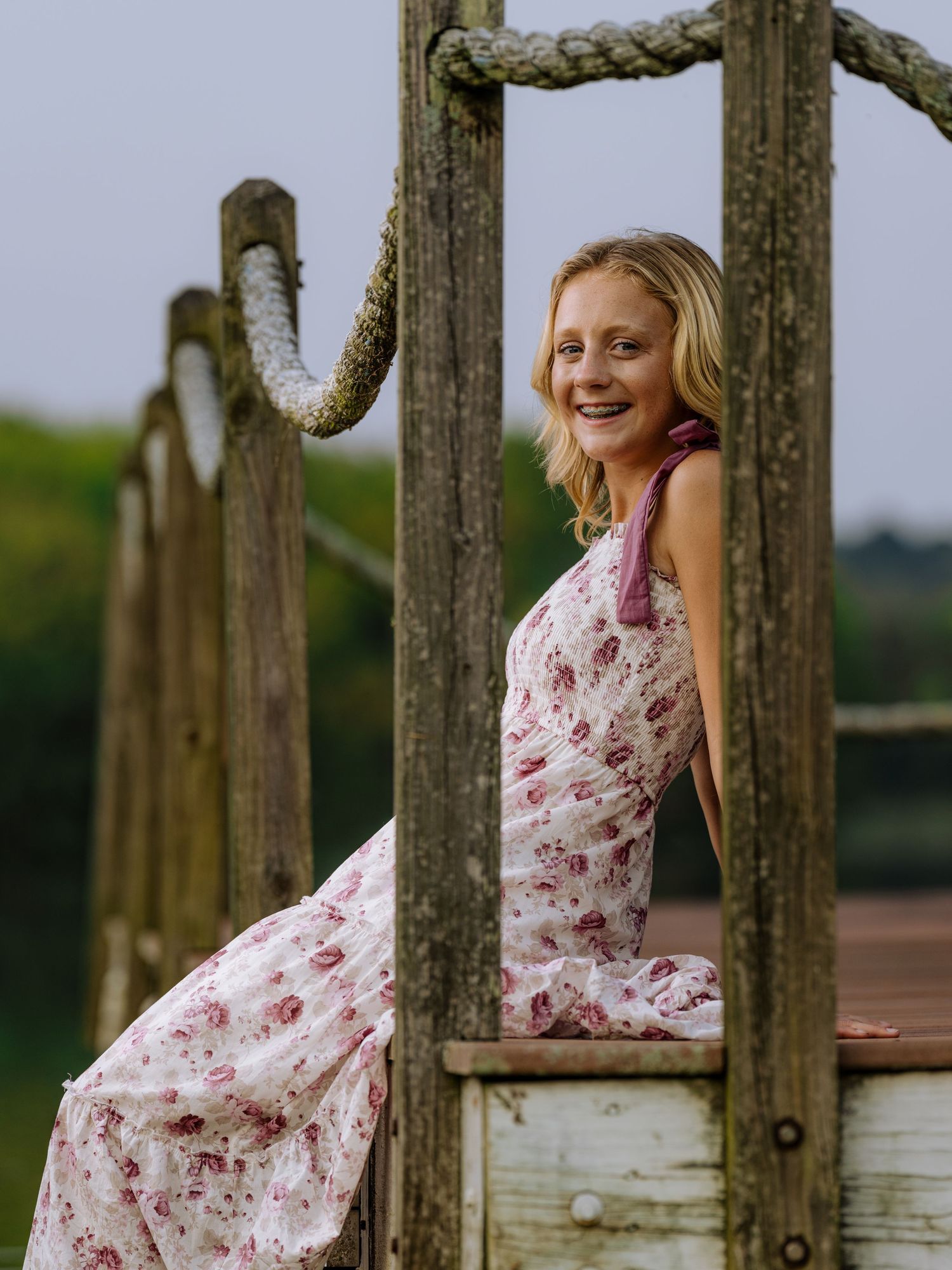Smiling blonde woman in floral dress posing on a rustic wooden rope bridge near a lake.