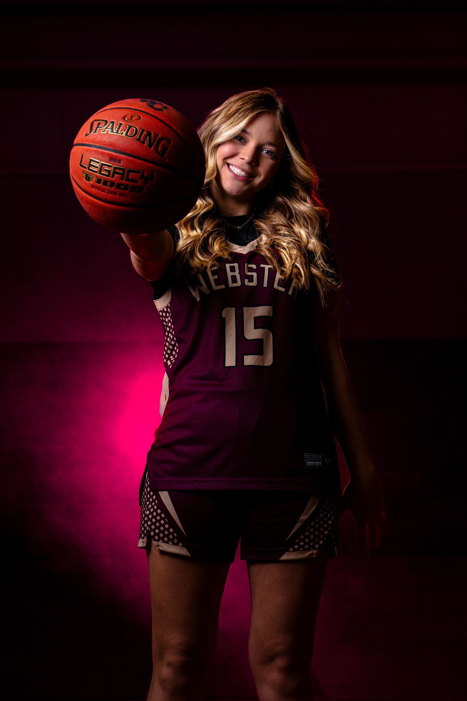 Basketball player in Webster #15 jersey poses with ball against dark background with dramatic pink lighting.