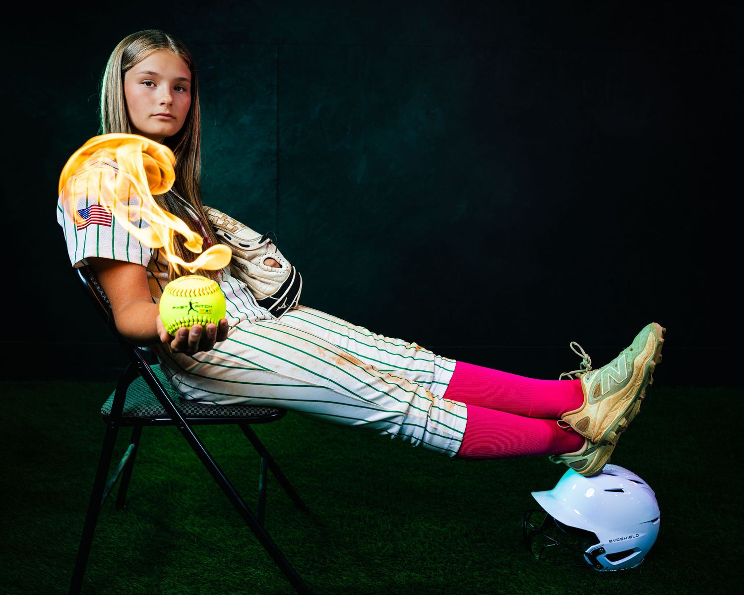 Young softball player in pinstripe uniform, pink socks, holding glove and ball, posed on chair with batting helmet nearby.