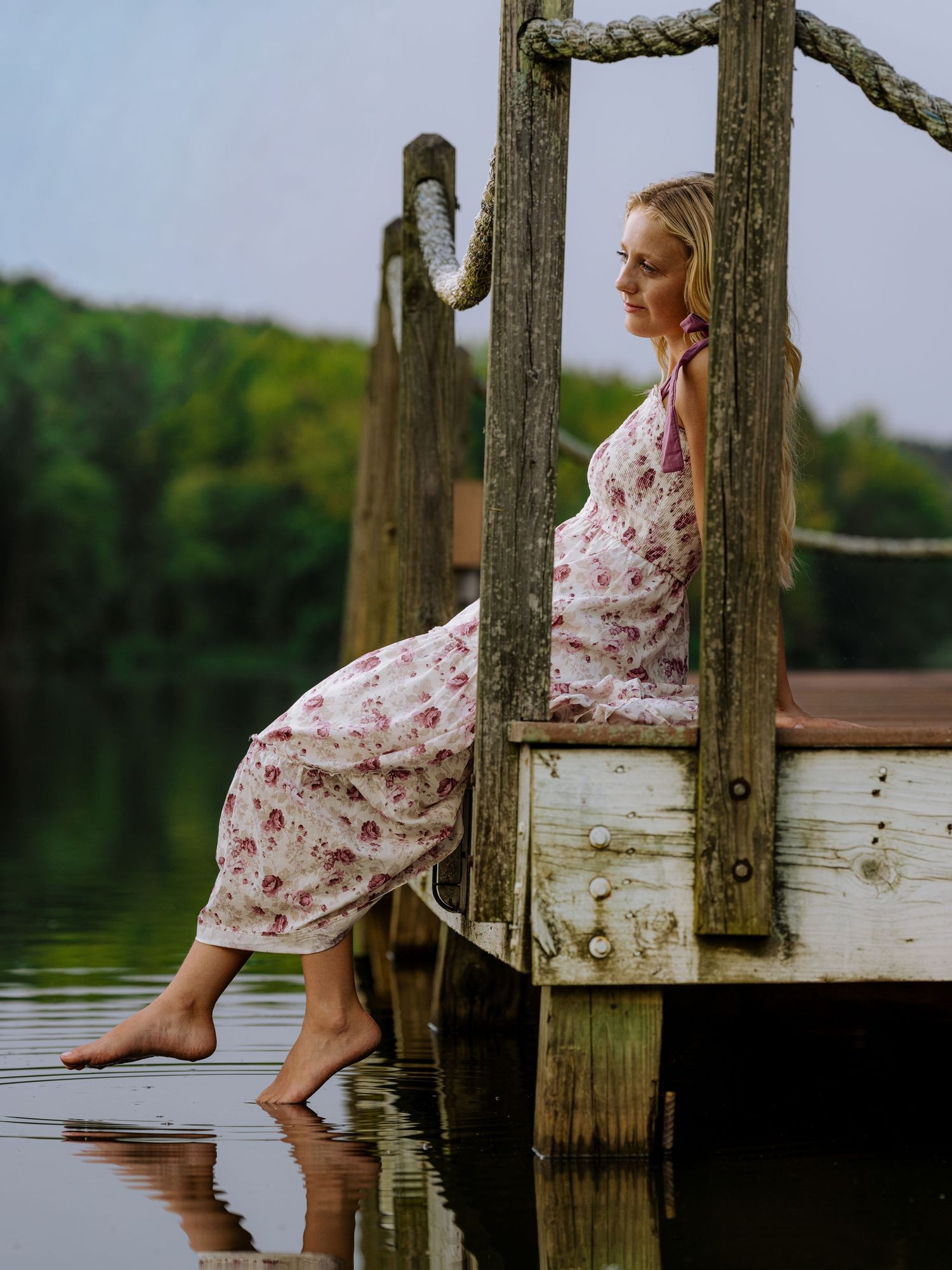 Barefoot pregnant woman in floral dress sitting on rustic wooden dock by calm lake surrounded by lush green trees.