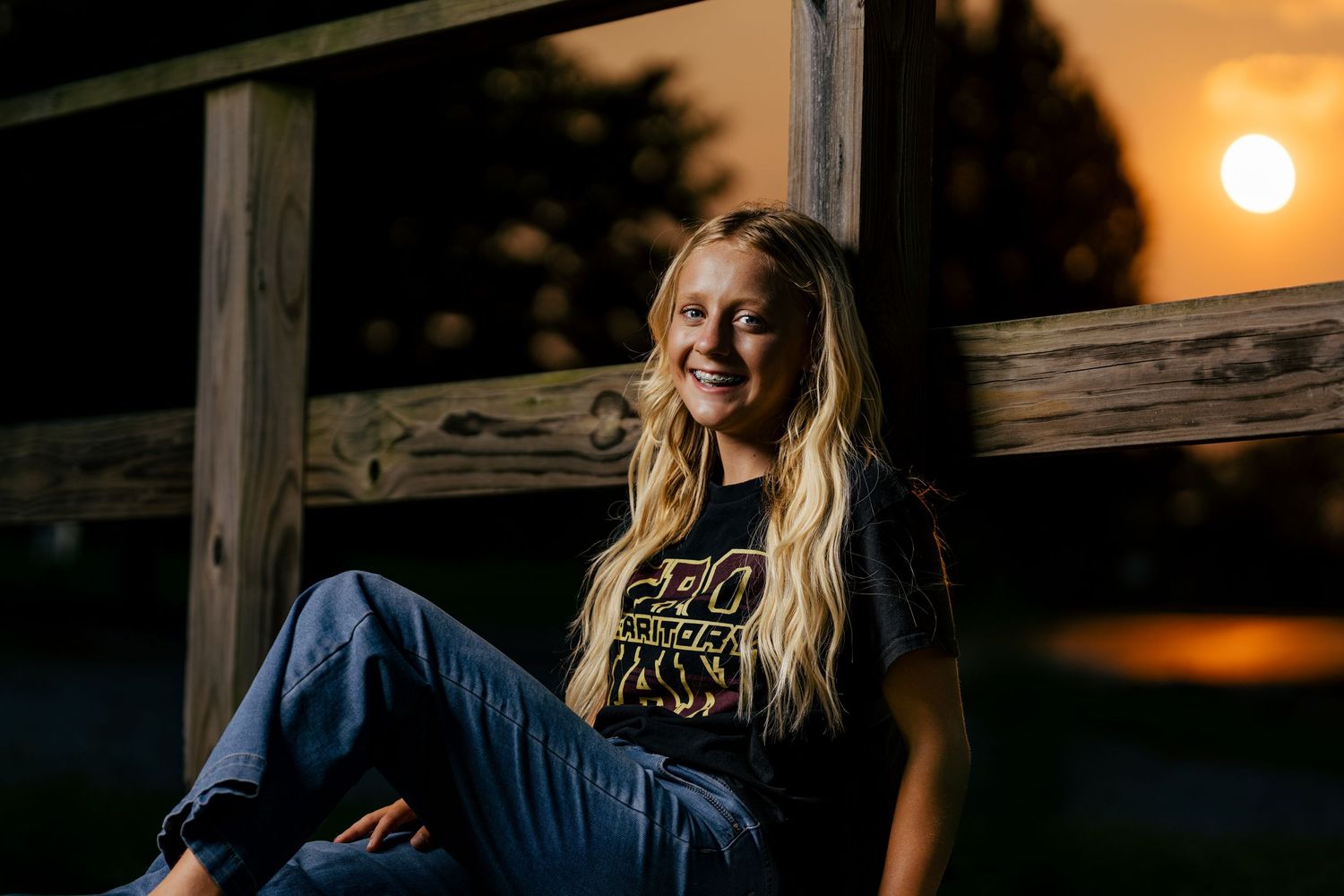 Young woman with long blonde hair smiling at sunset, sitting against a wooden fence in jeans and a black graphic tee.