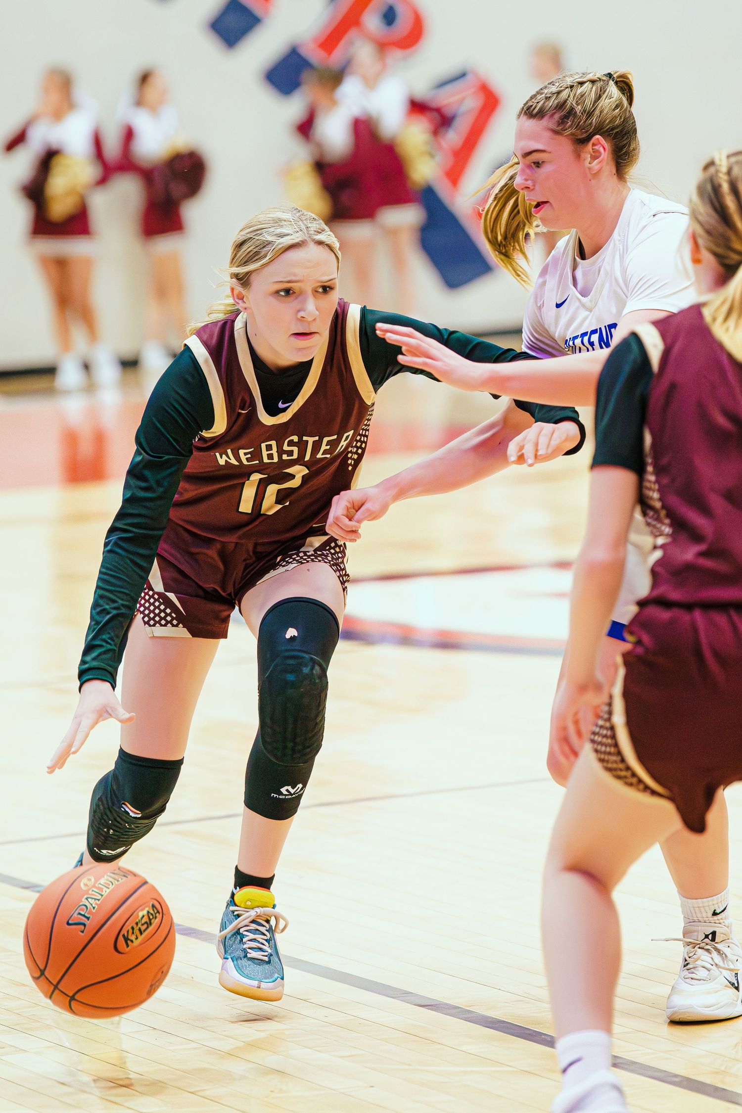 Female basketball player #12 in maroon Webster jersey dribbles past defender during indoor high school girls basketball game.