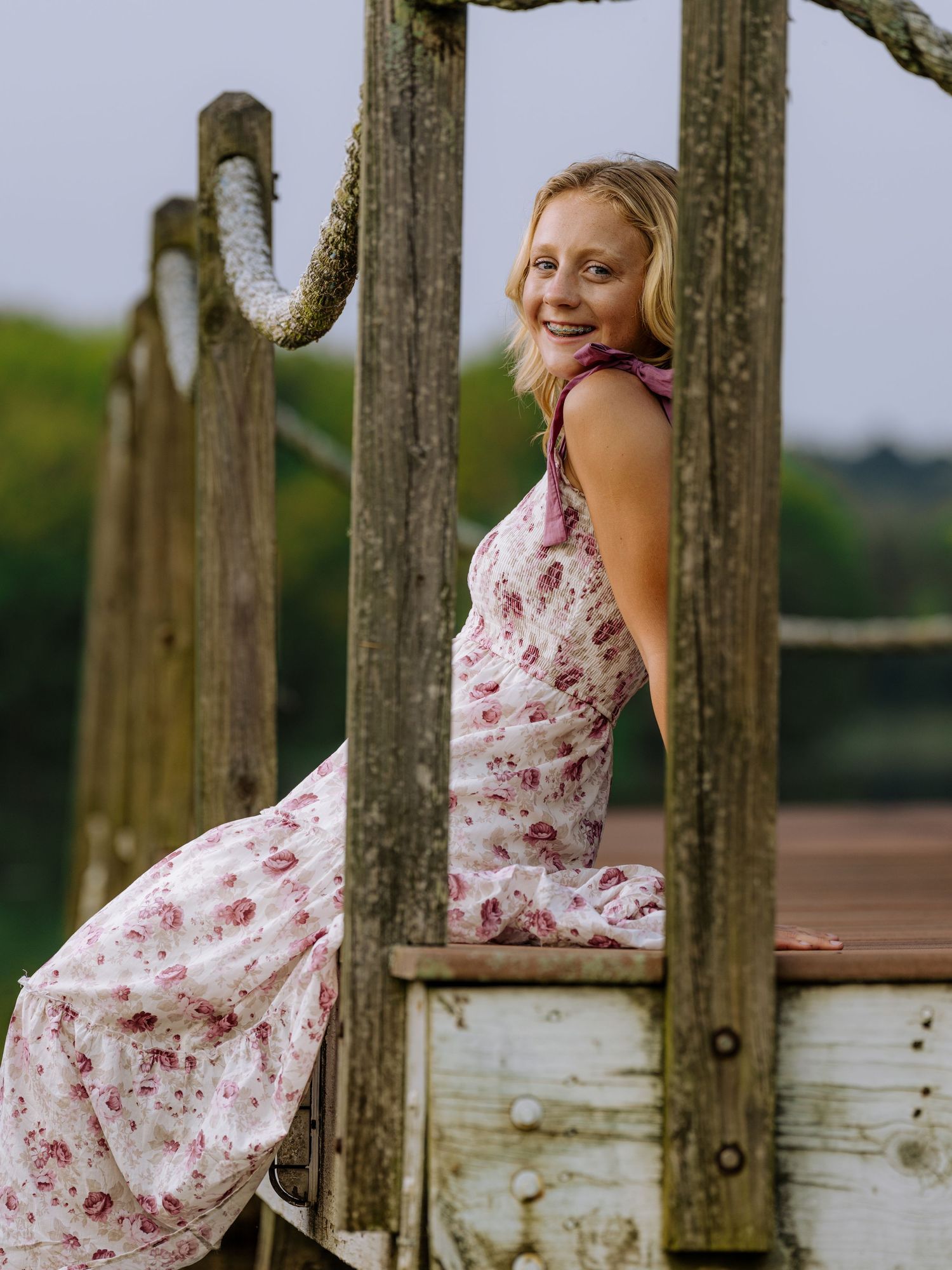 Young blonde woman in floral sundress smiling on rustic wooden dock bridge surrounded by greenery.
