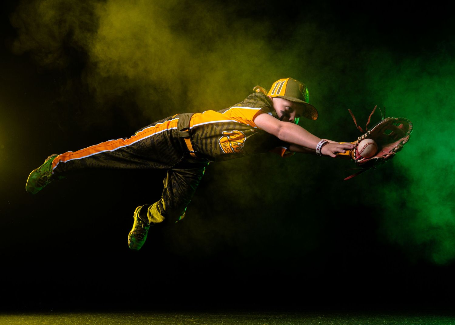 Baseball player in yellow uniform diving to catch a ball mid-air against a dramatic green and yellow smoke background.