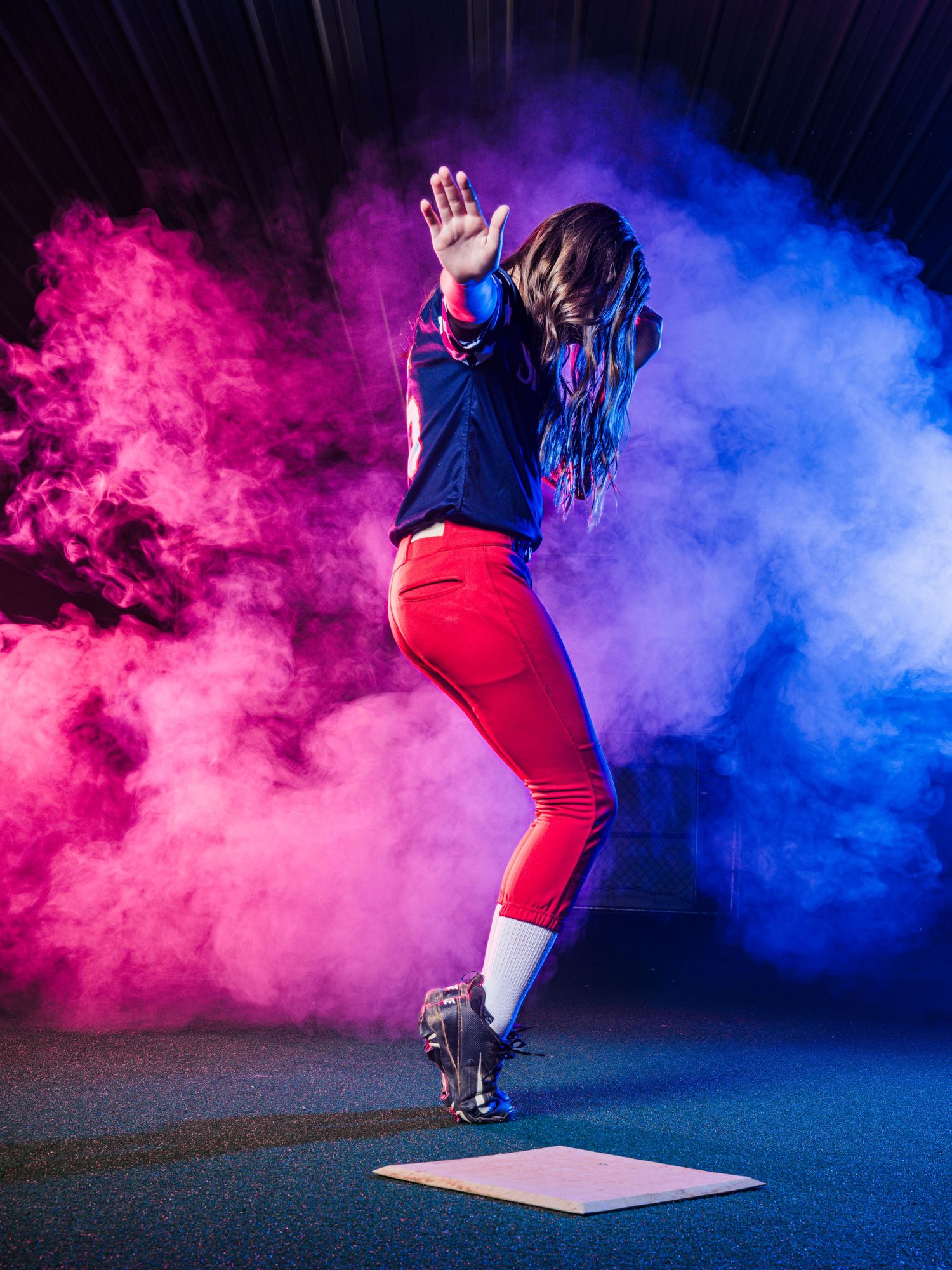 A female baseball player in red pants strikes a dynamic pose amid vibrant pink and blue smoke clouds on a dark background.