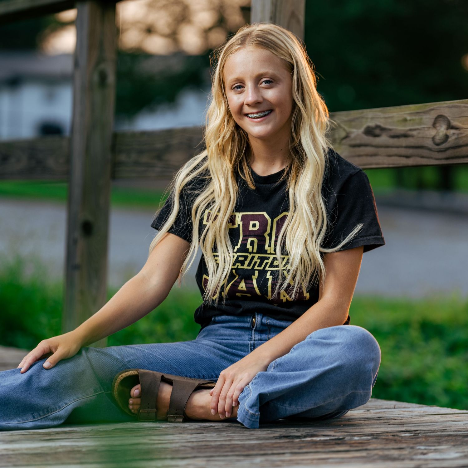 Smiling teenage girl with blonde hair sitting cross-legged on wooden deck wearing black college t-shirt and jeans.