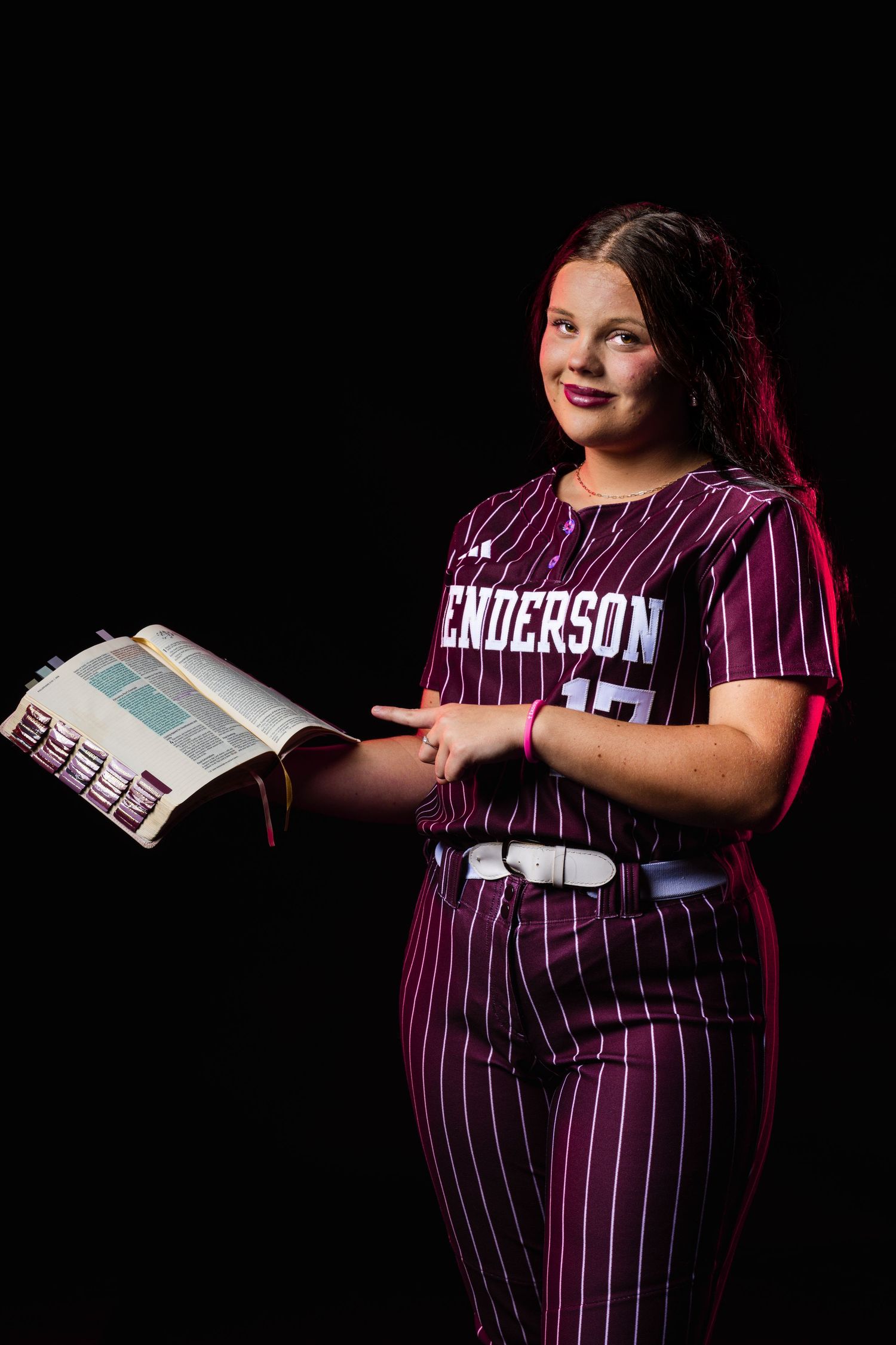 A young softball player in a maroon Henderson pinstripe uniform holds an open book, posing against a black background.