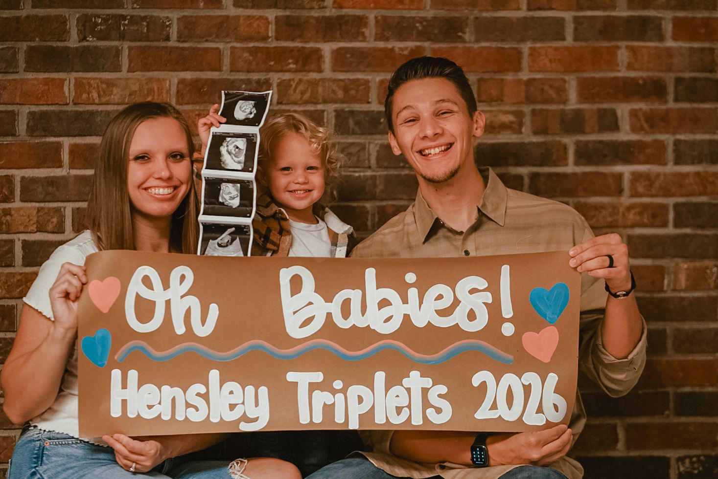 Family pregnancy announcement with wooden signs against brick wall backdrop.