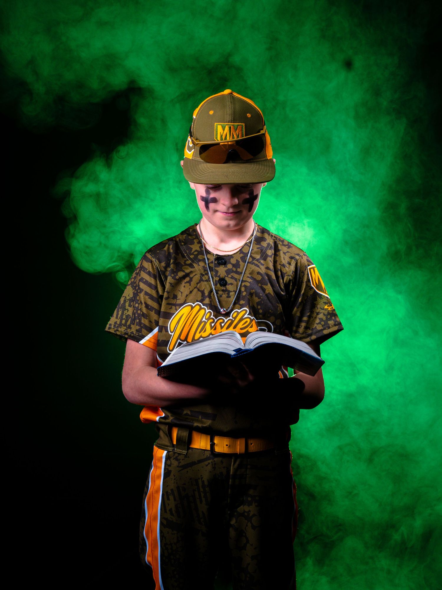 Young baseball player in Missiles uniform and camo hat reads a book against dramatic green smoke background.