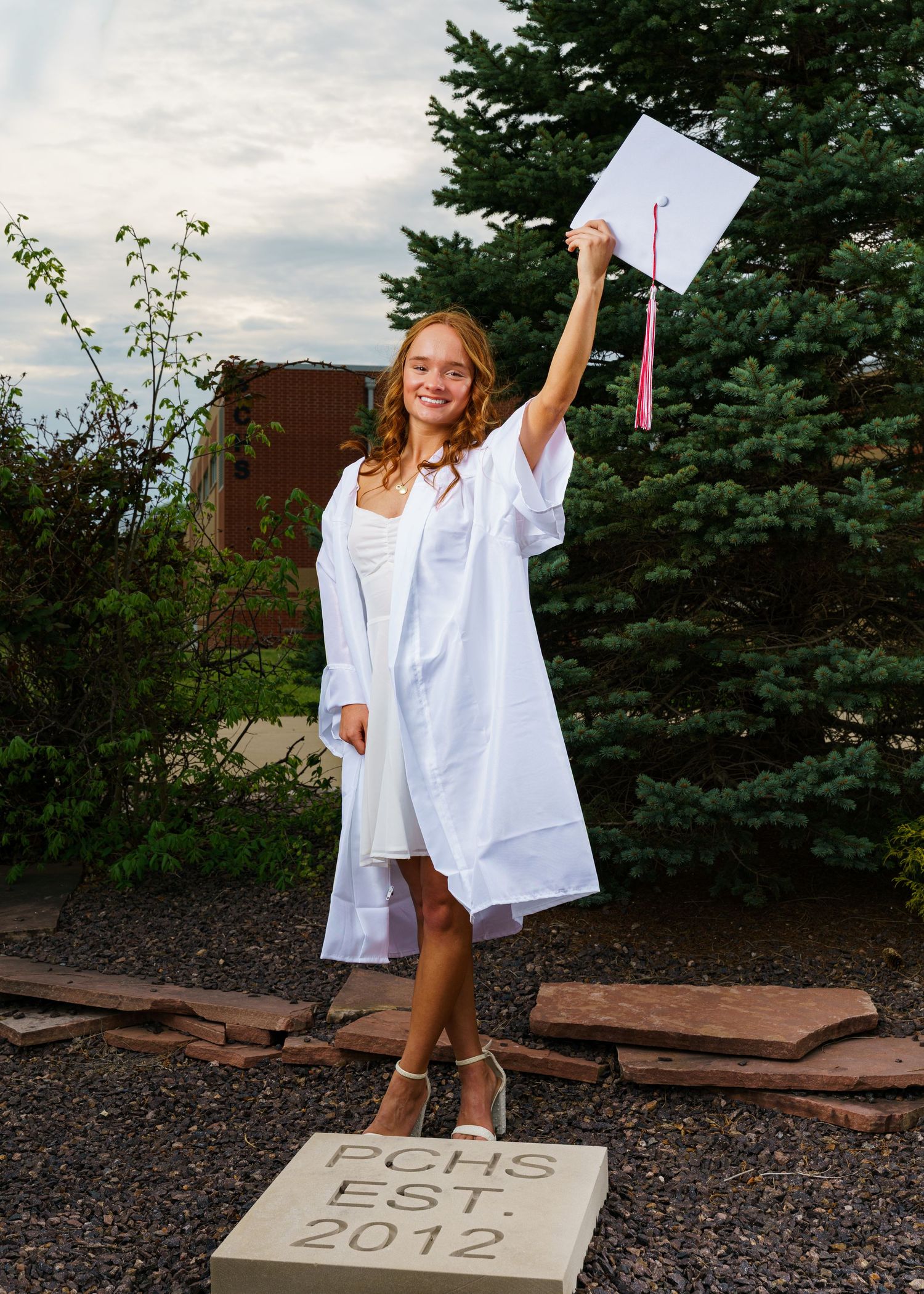 Smiling graduate in white cap and gown holds diploma aloft outdoors near PCHS Est. 2012 stone marker.