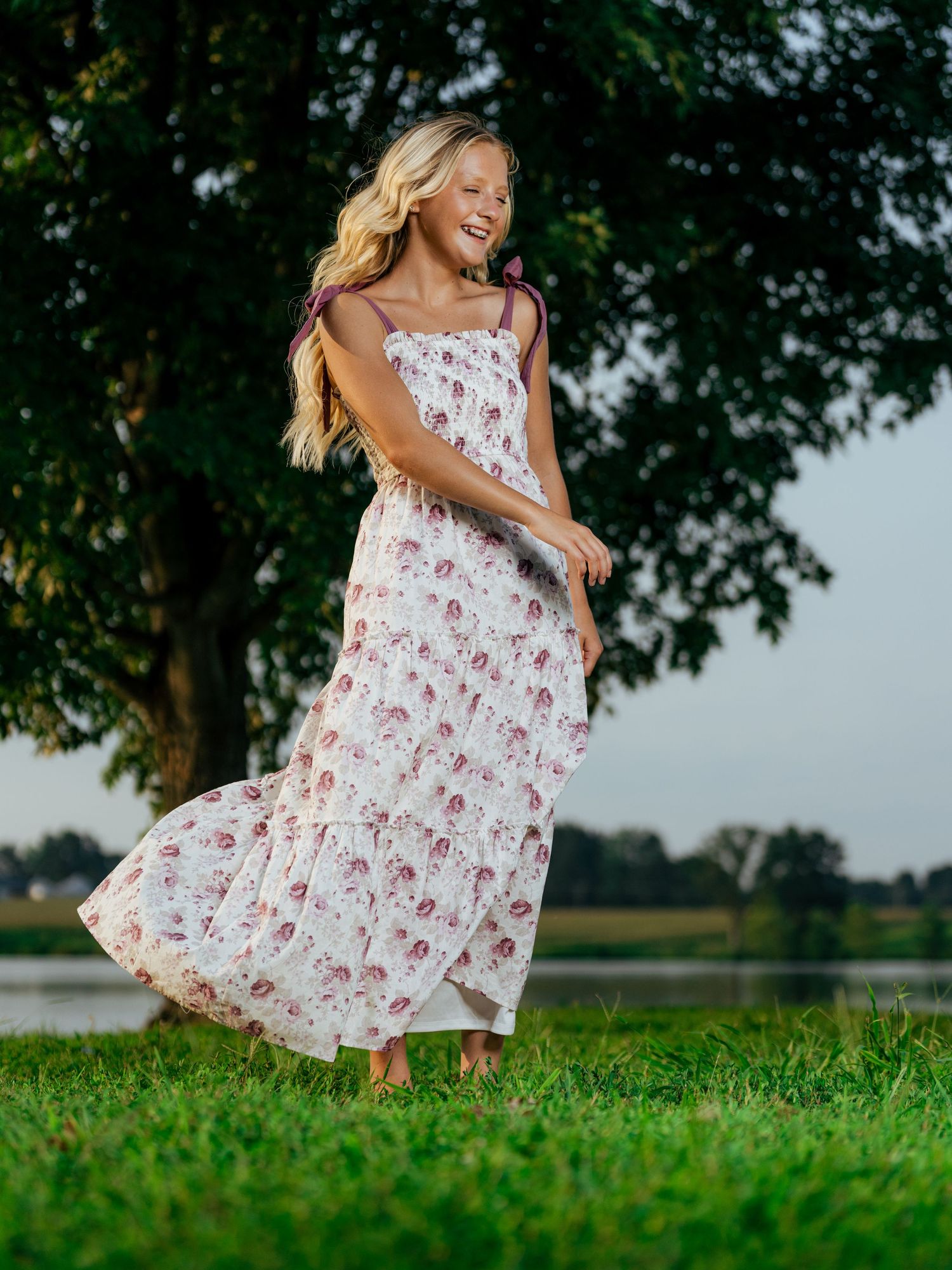 Young blonde girl twirling in a white floral maxi dress on green grass near a tree.