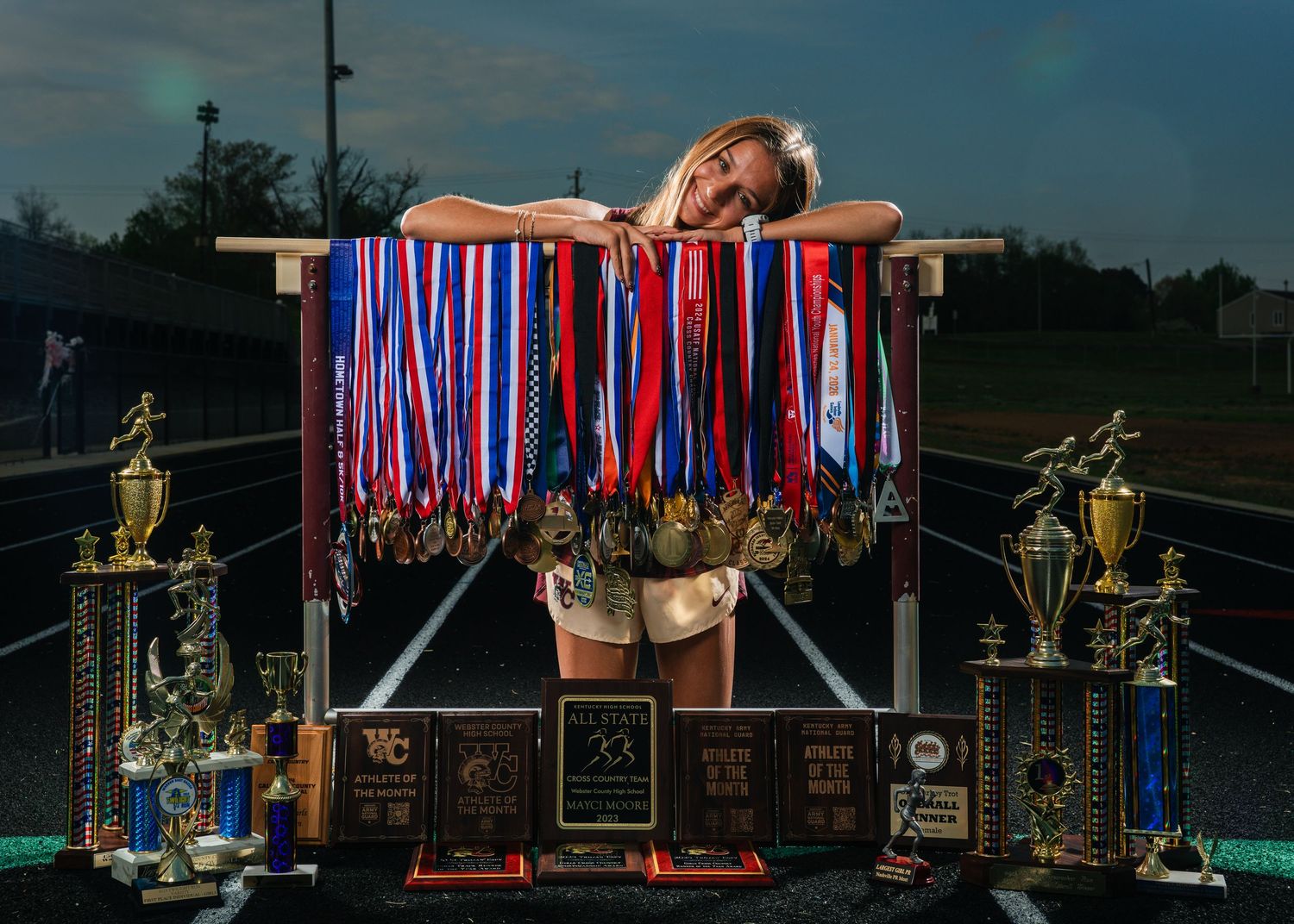 Young female athlete poses with numerous medals, ribbons, and trophies displayed on a track field at dusk.