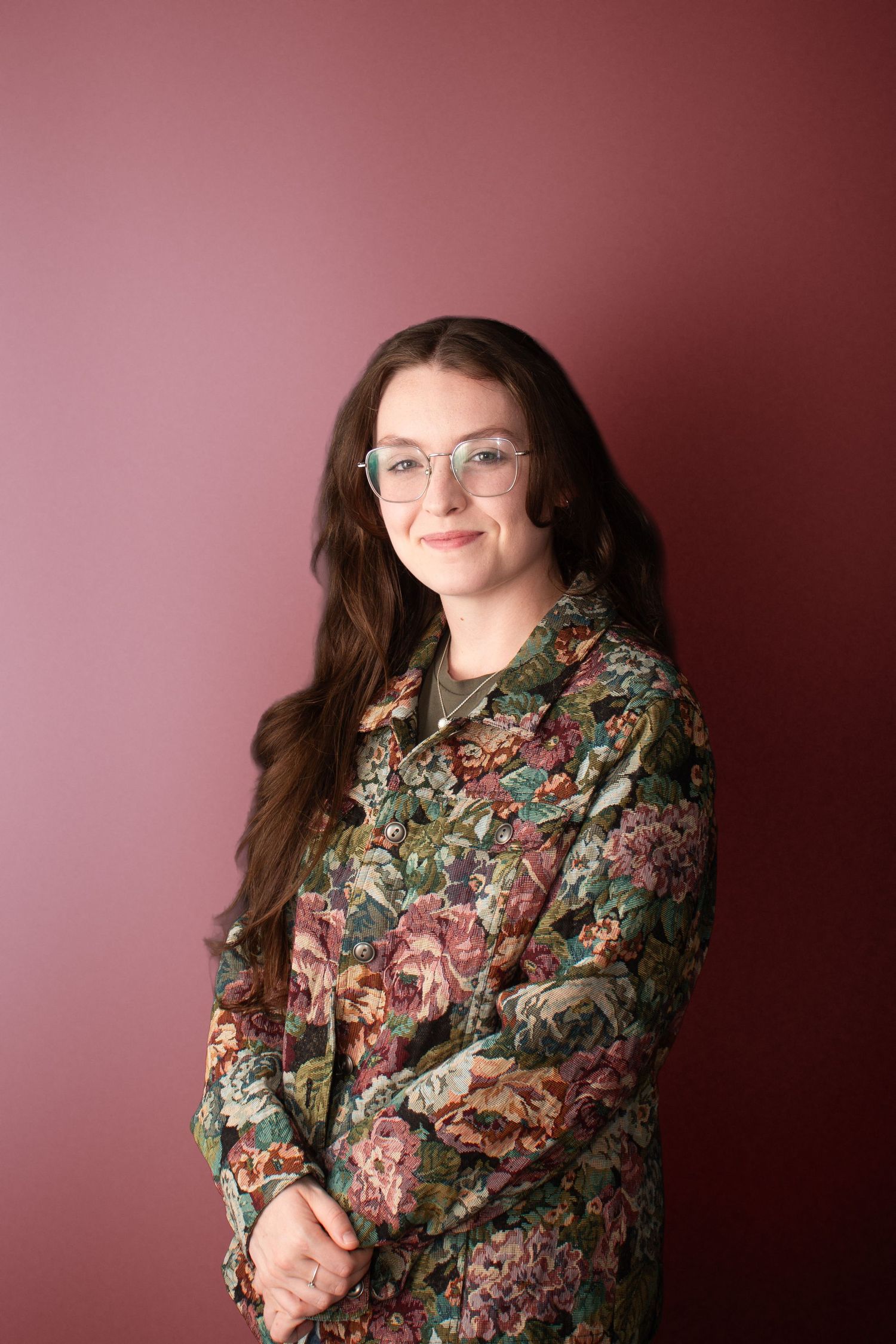A smiling young woman wearing glasses and a floral jacket poses against a mauve background in a portrait.
